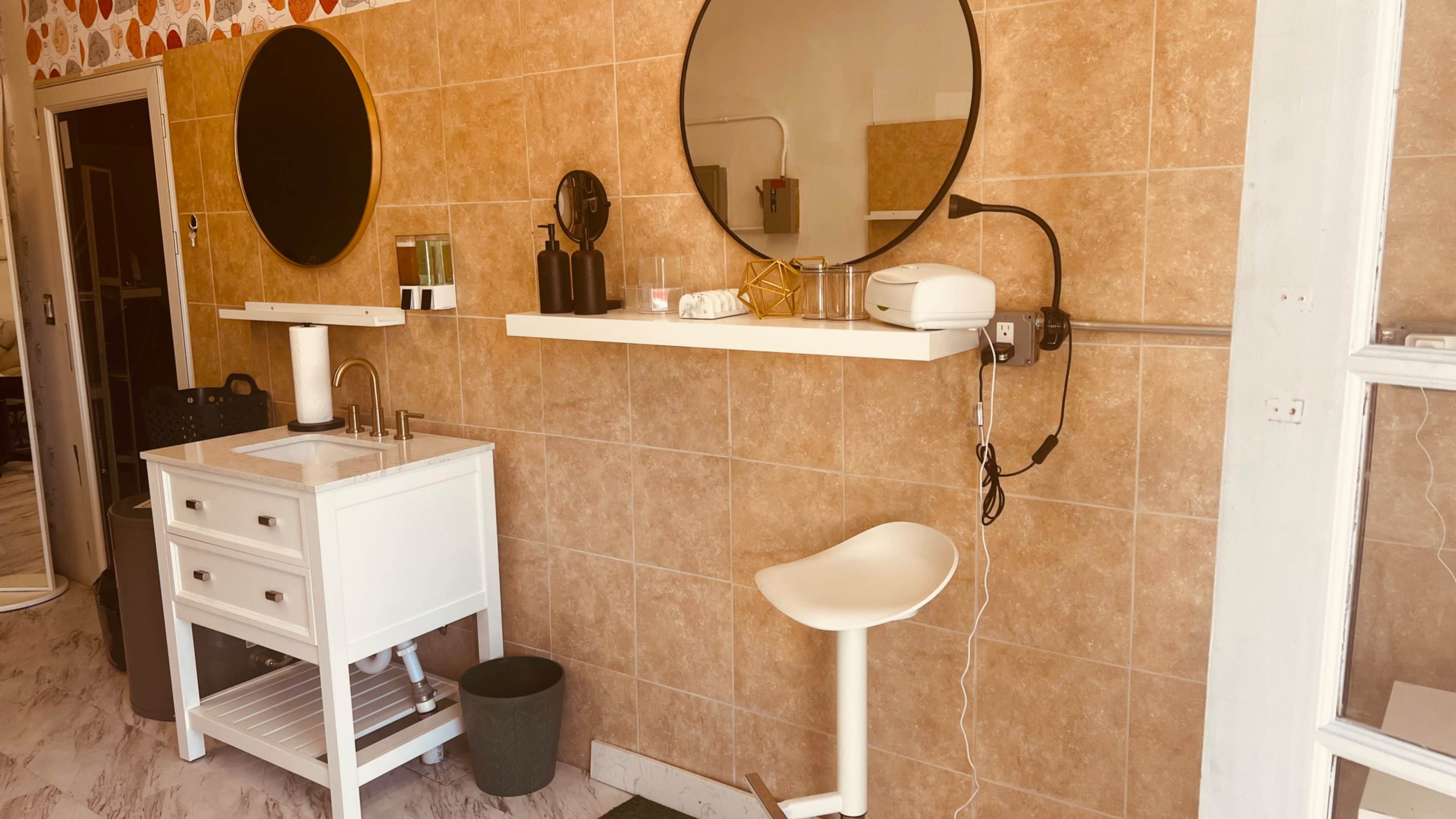The image shows a minimalist salon interior featuring two round mirrors above a white countertop with a sink, complemented by a bar stool and a small waste bin.
