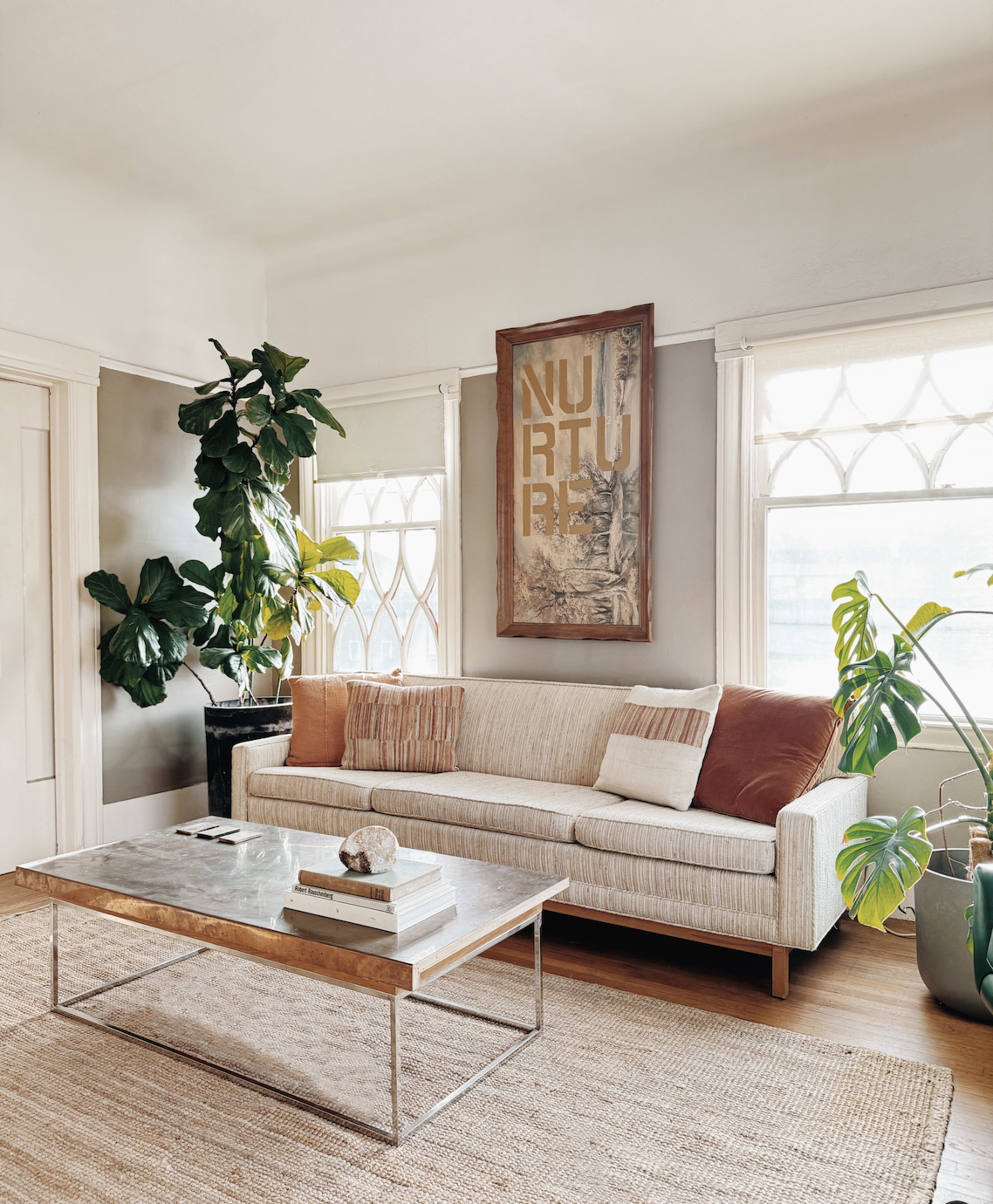 A modern living room features a light-colored sofa, a glass-top coffee table, and potted plants near large windows.