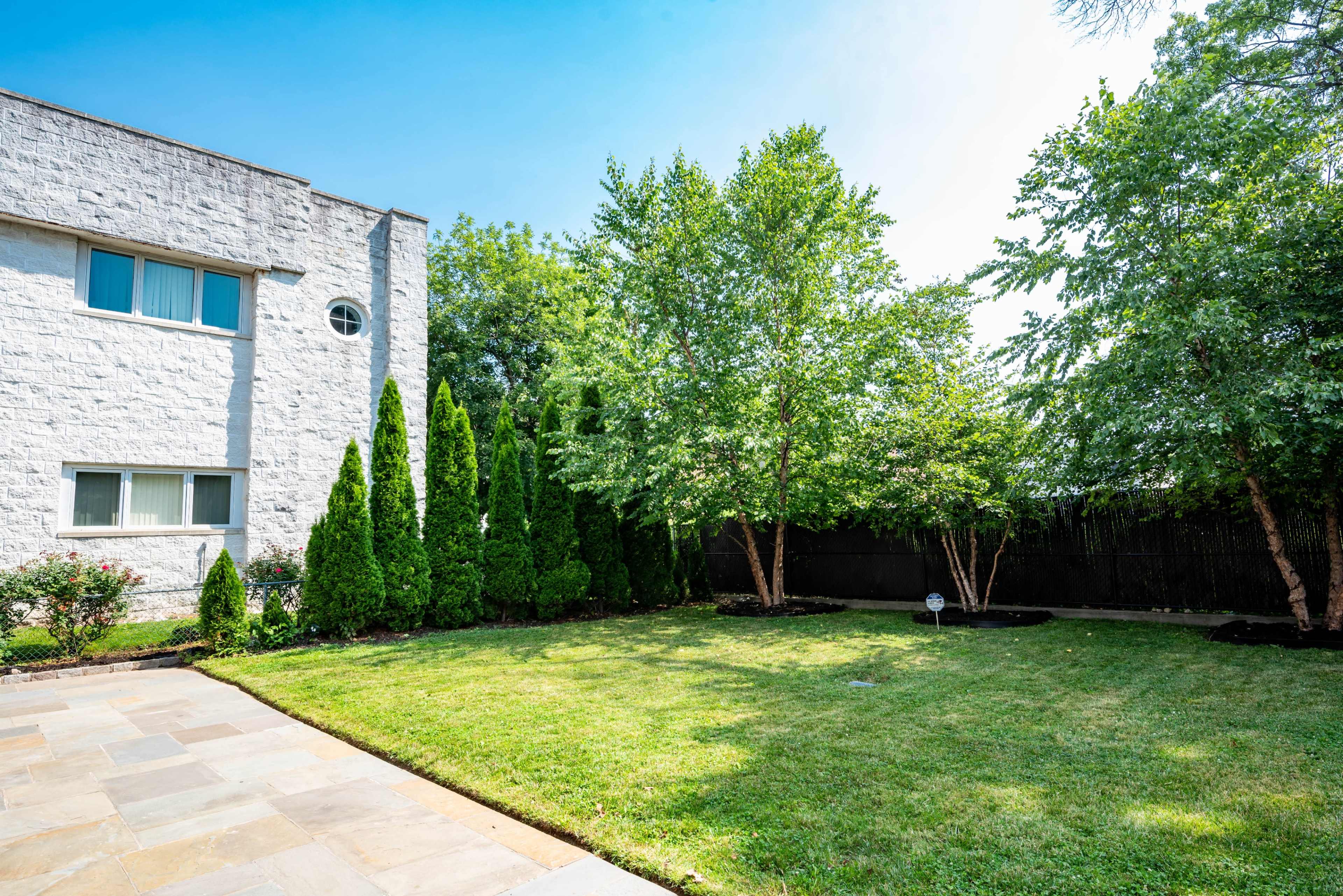 The image shows a well-maintained backyard with a neatly trimmed lawn, bordered by several trees and shrubs, and a stone patio adjacent to the house.