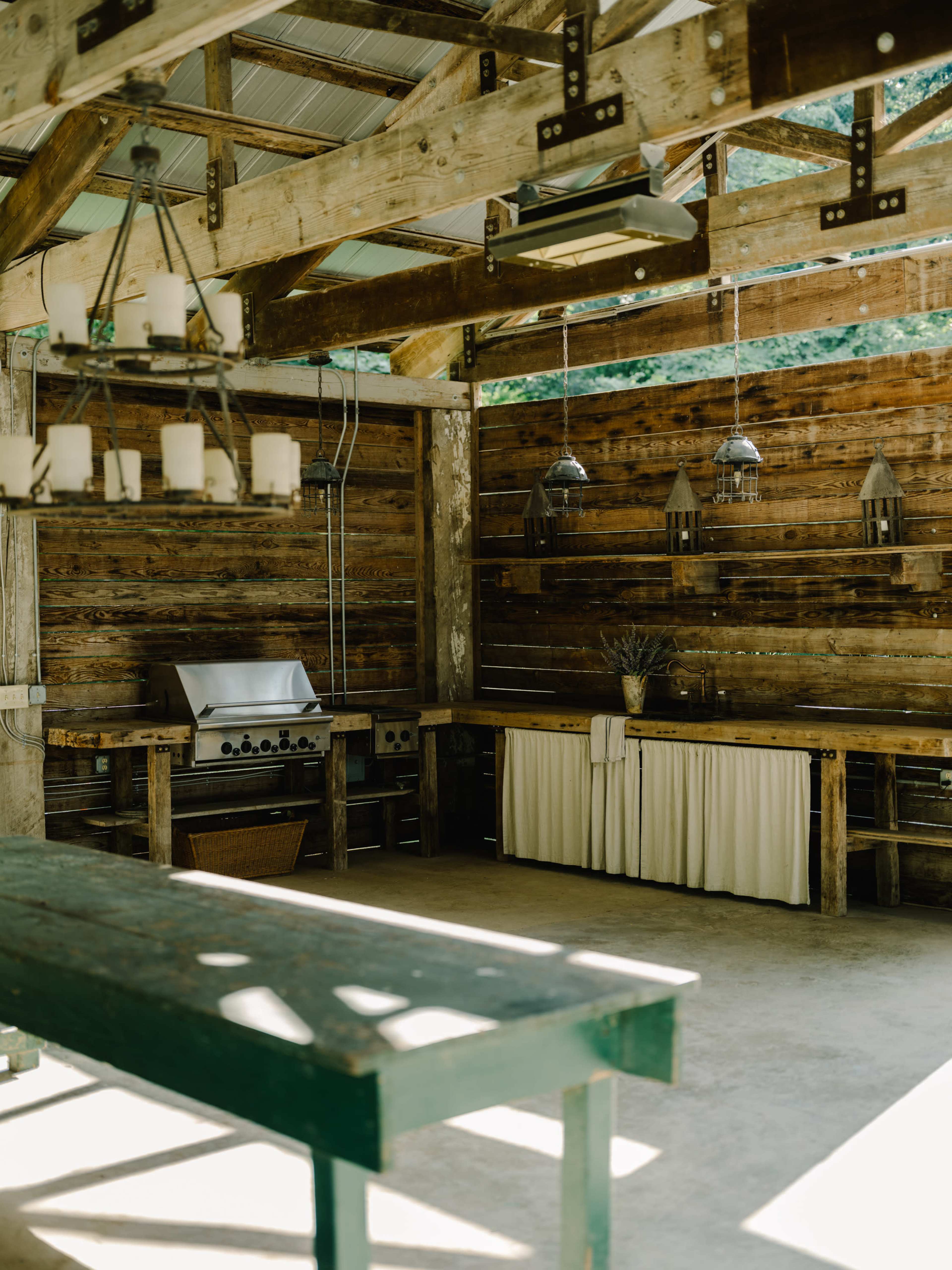 The image shows a rustic outdoor kitchen with wooden walls, a stainless steel grill, and a large table made of wood.