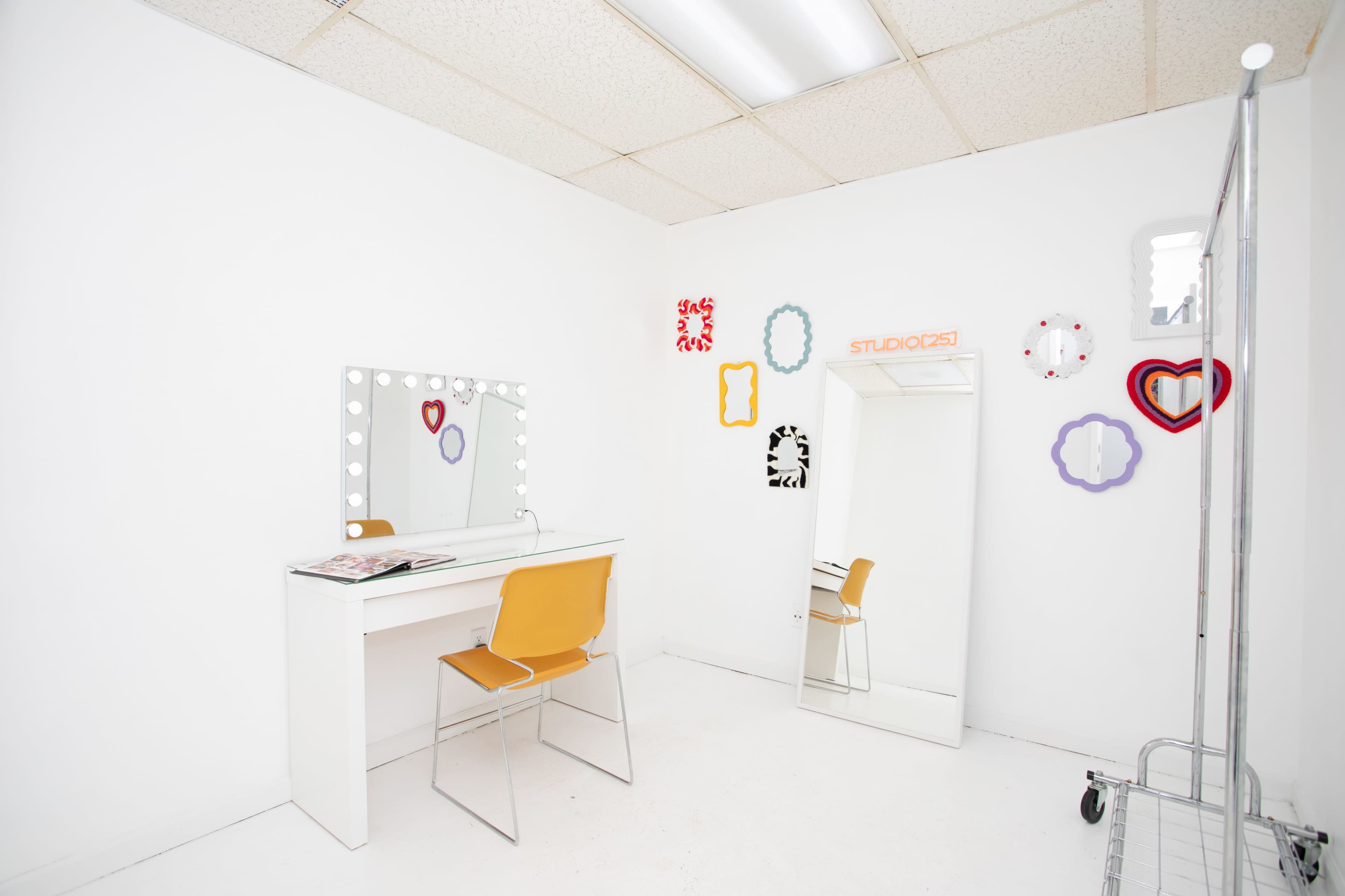 The image shows a bright, white room featuring a vanity with a mirror and lights, a chair, a full-length mirror, and decorative frames on the walls.