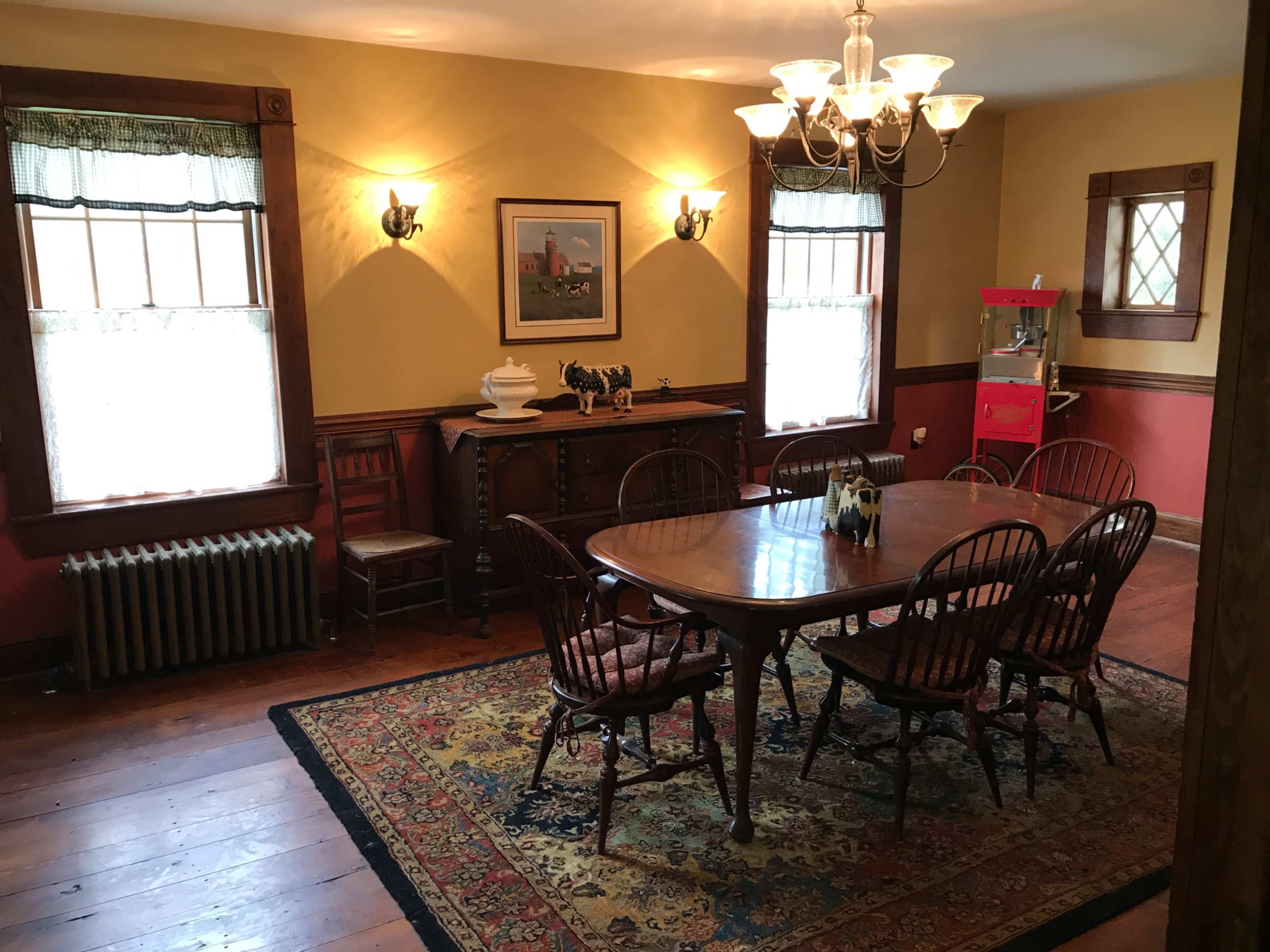 The image shows a dining room with a wooden table surrounded by chairs, a colorful rug on the floor, and a vintage popcorn machine in the corner.