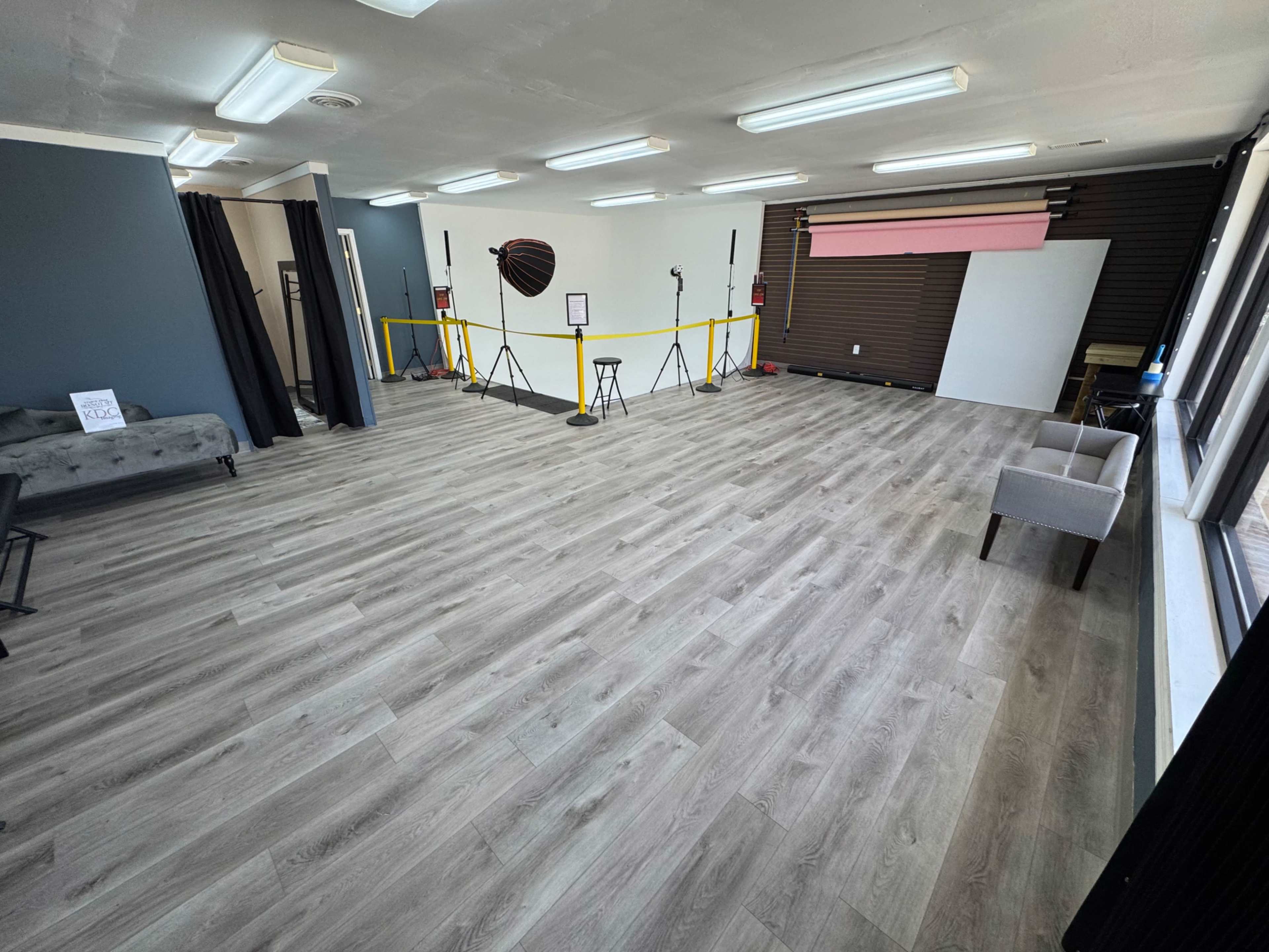 The image shows a spacious, empty room with light wooden flooring, equipped with photography lighting setups and a backdrop.