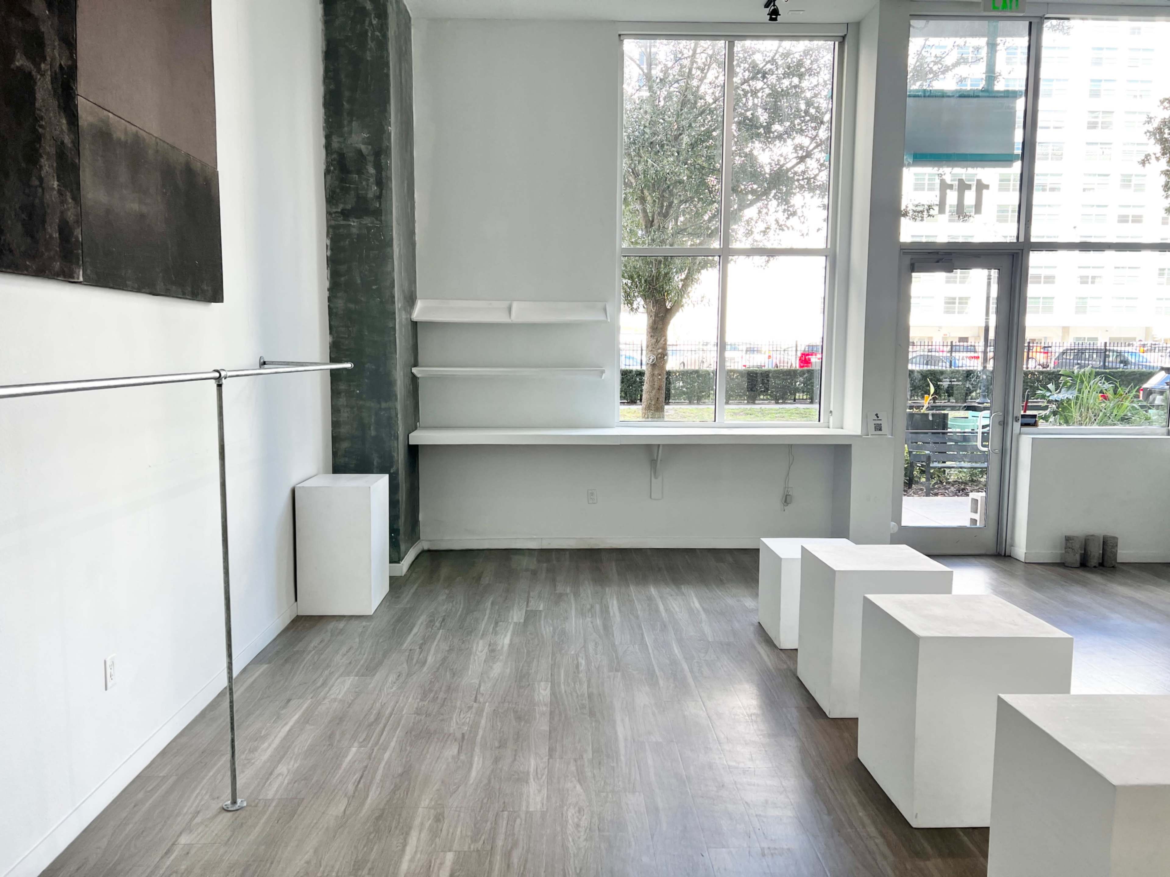 A minimalist interior space featuring a clothing rack, three white cubic stools, and a shelf against a wall with large windows allowing natural light.