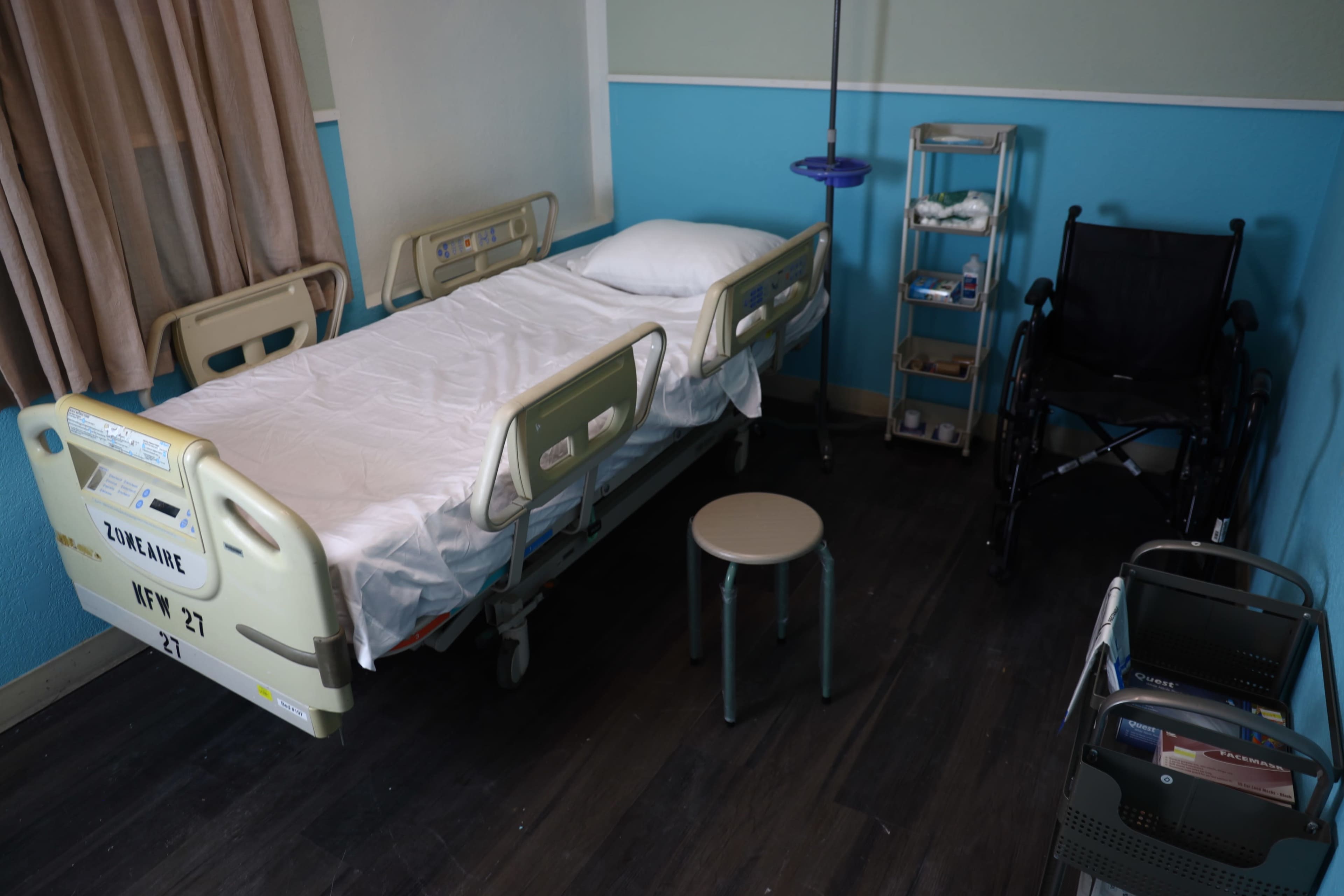 A hospital room features an unoccupied bed, a bedside stool, a wheelchair, and a mobile storage cart.