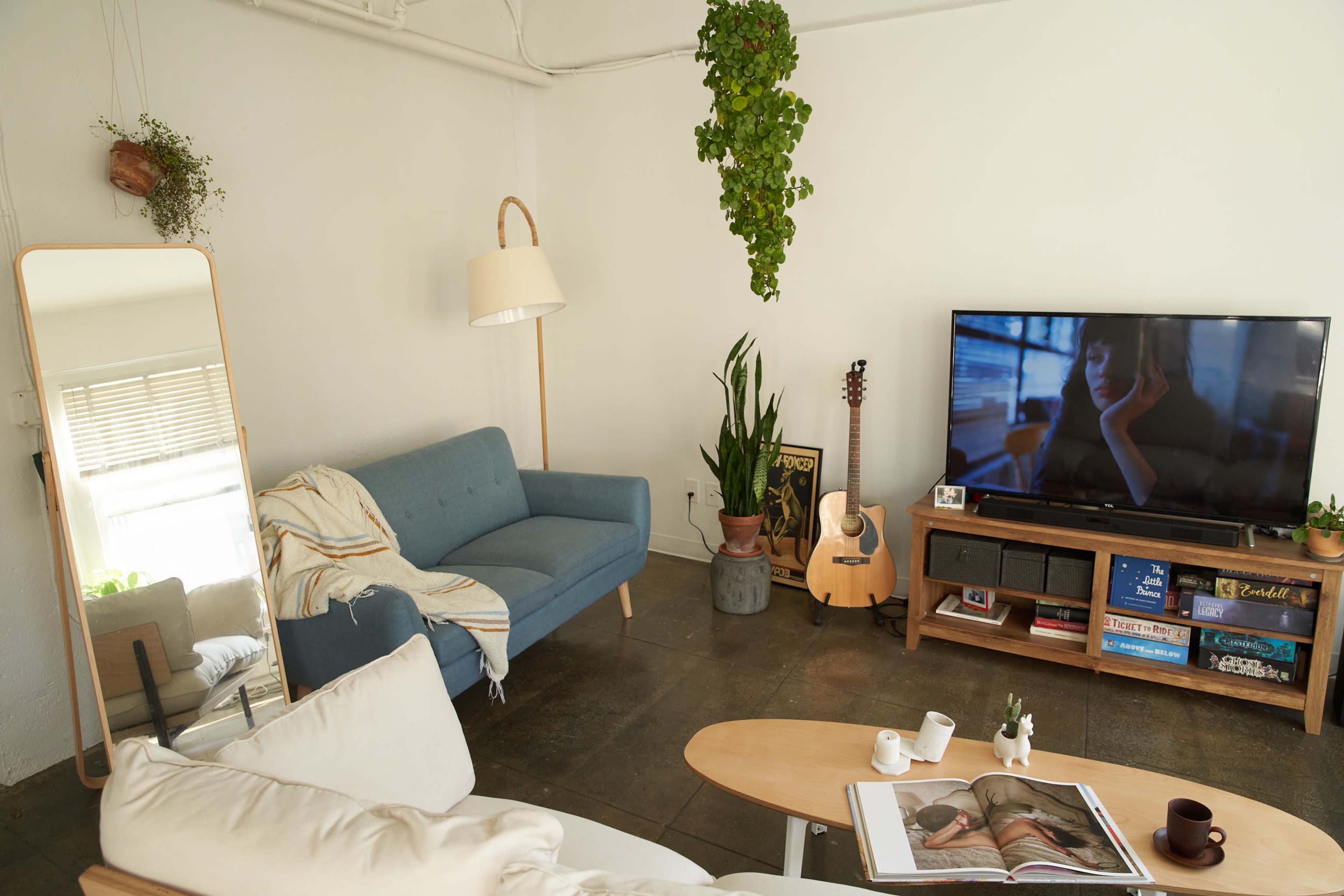 A cozy living room features a blue sofa, a large mirror, a guitar, and a television displaying a scene, all accented by plants and wooden furniture.