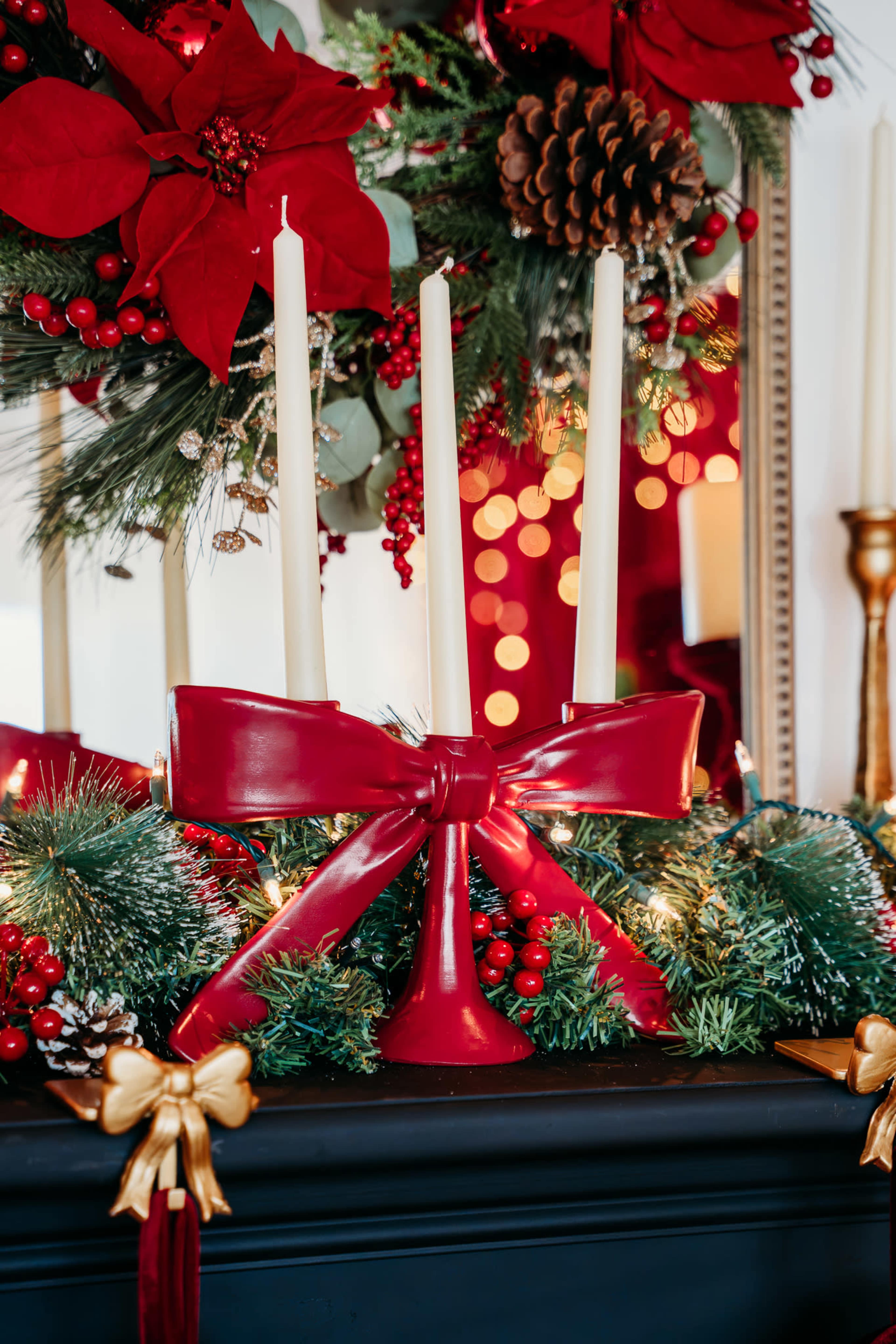 A centerpiece of three white candles tied with a red bow is displayed on a mantle adorned with greenery, red berries, and festive decorations.