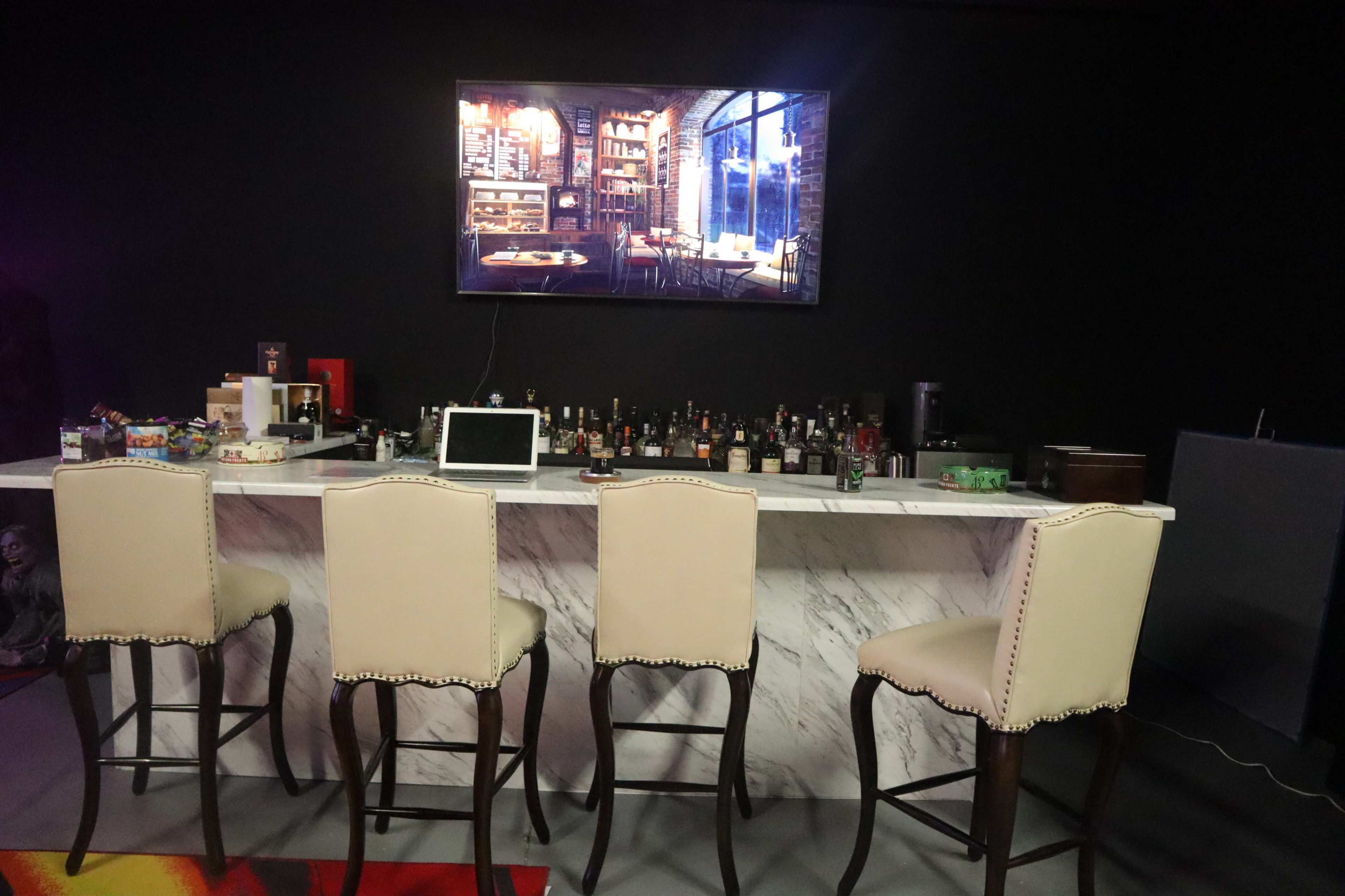 A bar with four high stools and a wall-mounted television displaying an image of a library-like interior, featuring various bottles of liquor on the counter.