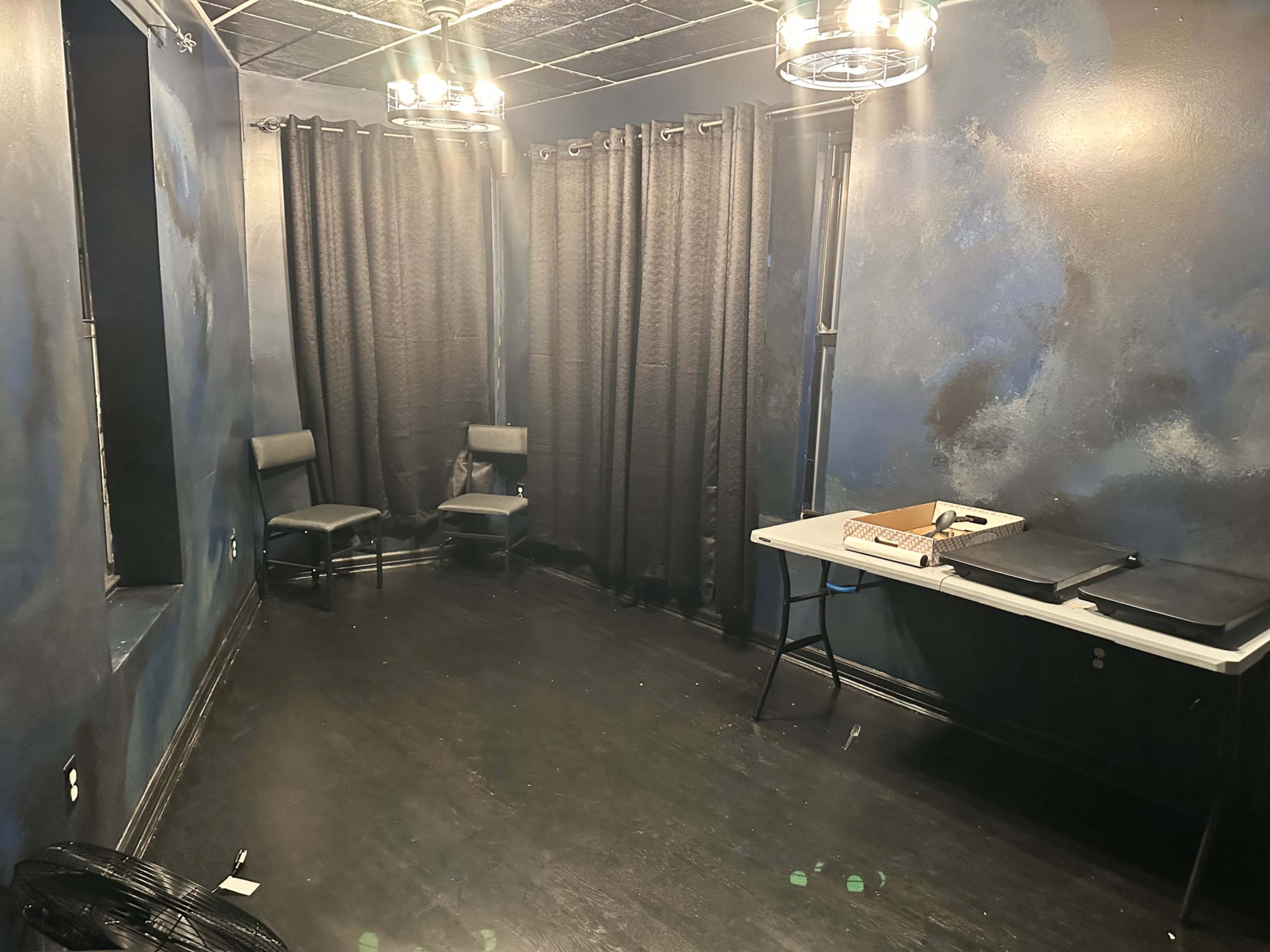 The room features two chairs, a table with folders, and black curtains covering the windows, all against a darkly painted wall.