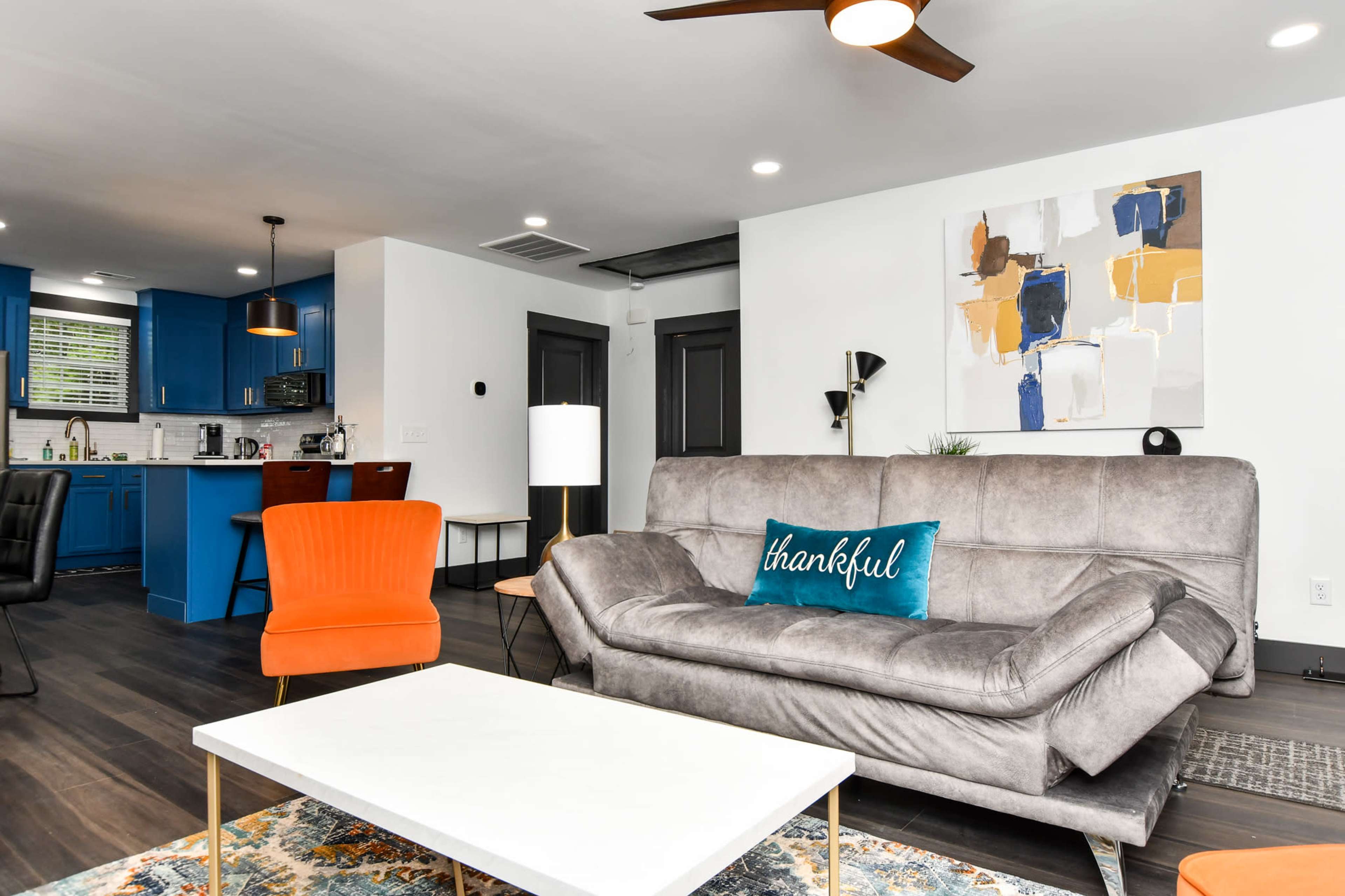The living area features a gray sectional sofa with a "thankful" pillow, a white coffee table, and vibrant orange chairs, accented by a blue kitchen in the background.