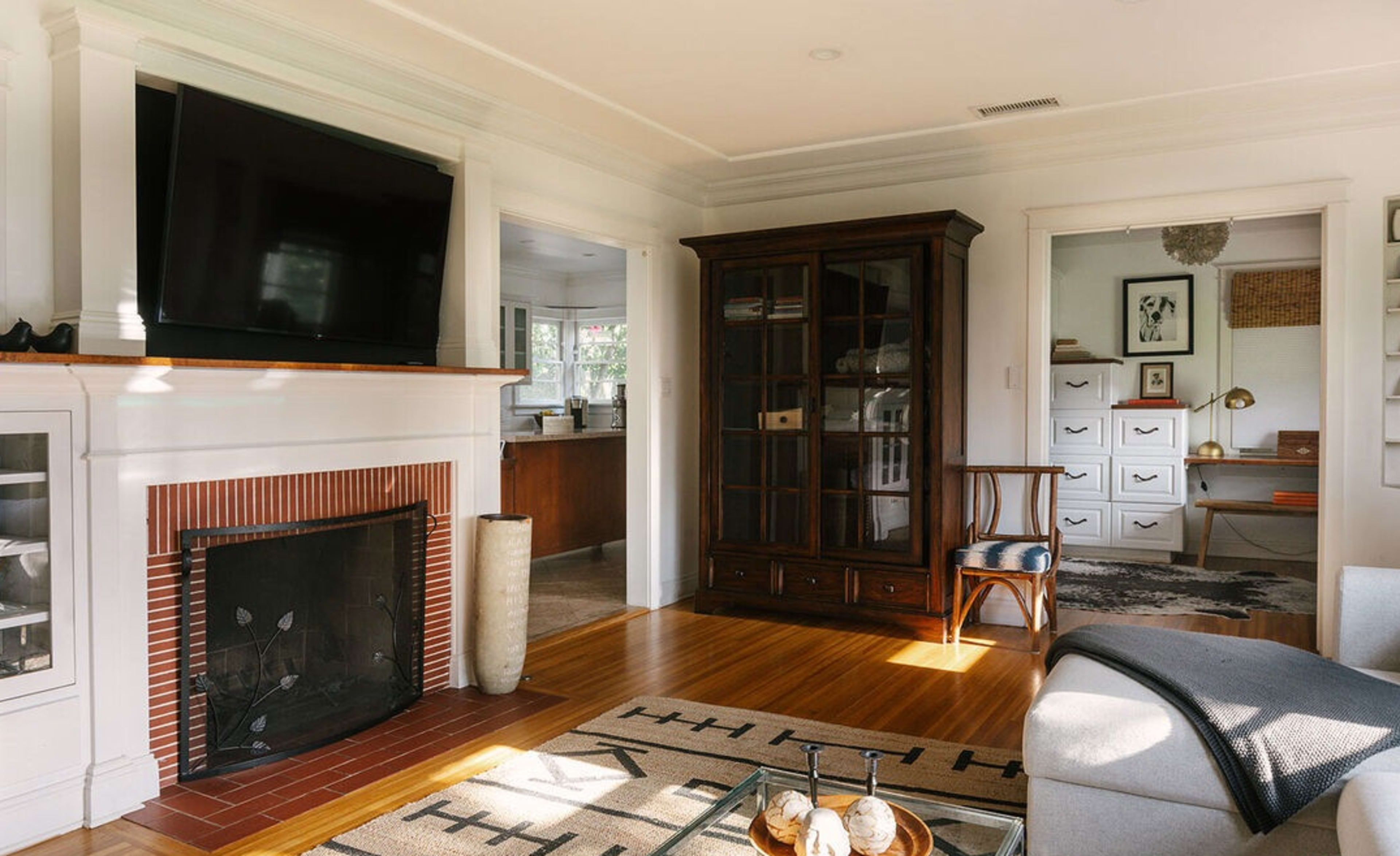 The image shows a cozy living room with a fireplace, a television mounted above it, and an adjacent room visible through an open doorway.