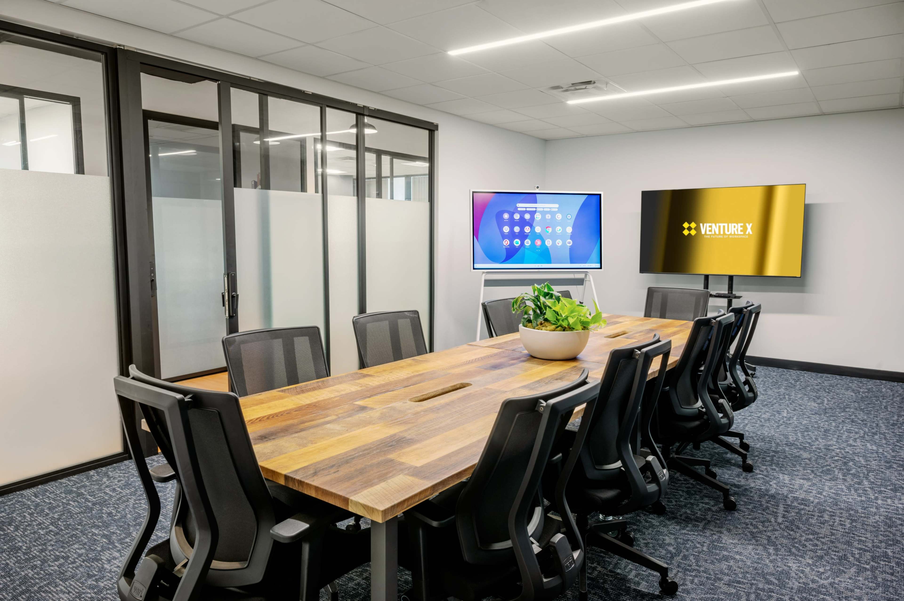 A modern conference room features a large wooden table surrounded by black office chairs, with screens displaying digital content on opposite walls.