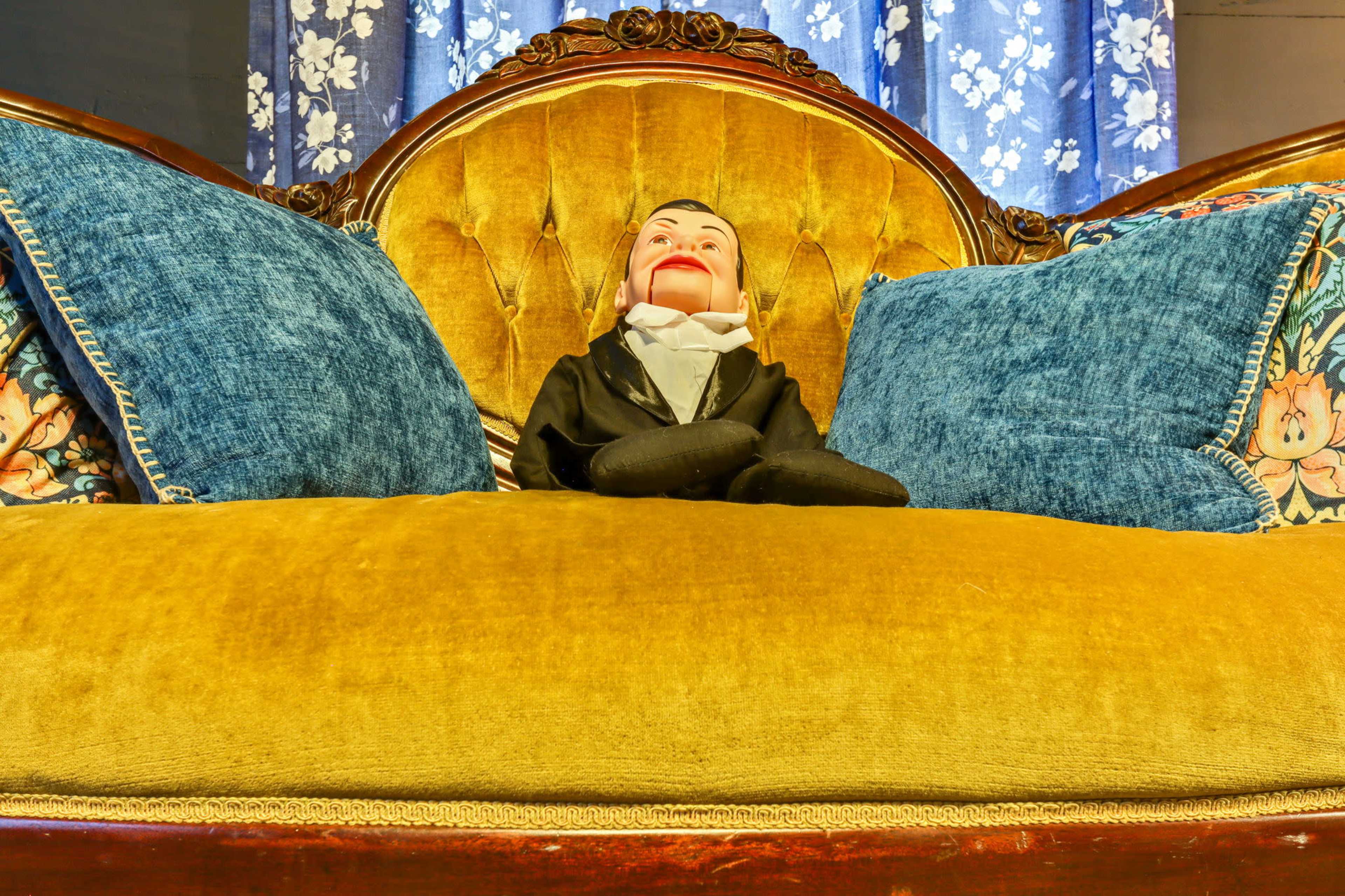 A vintage couch with a gold upholstery features two blue cushions and a ventriloquist dummy dressed in a suit sitting in the middle.