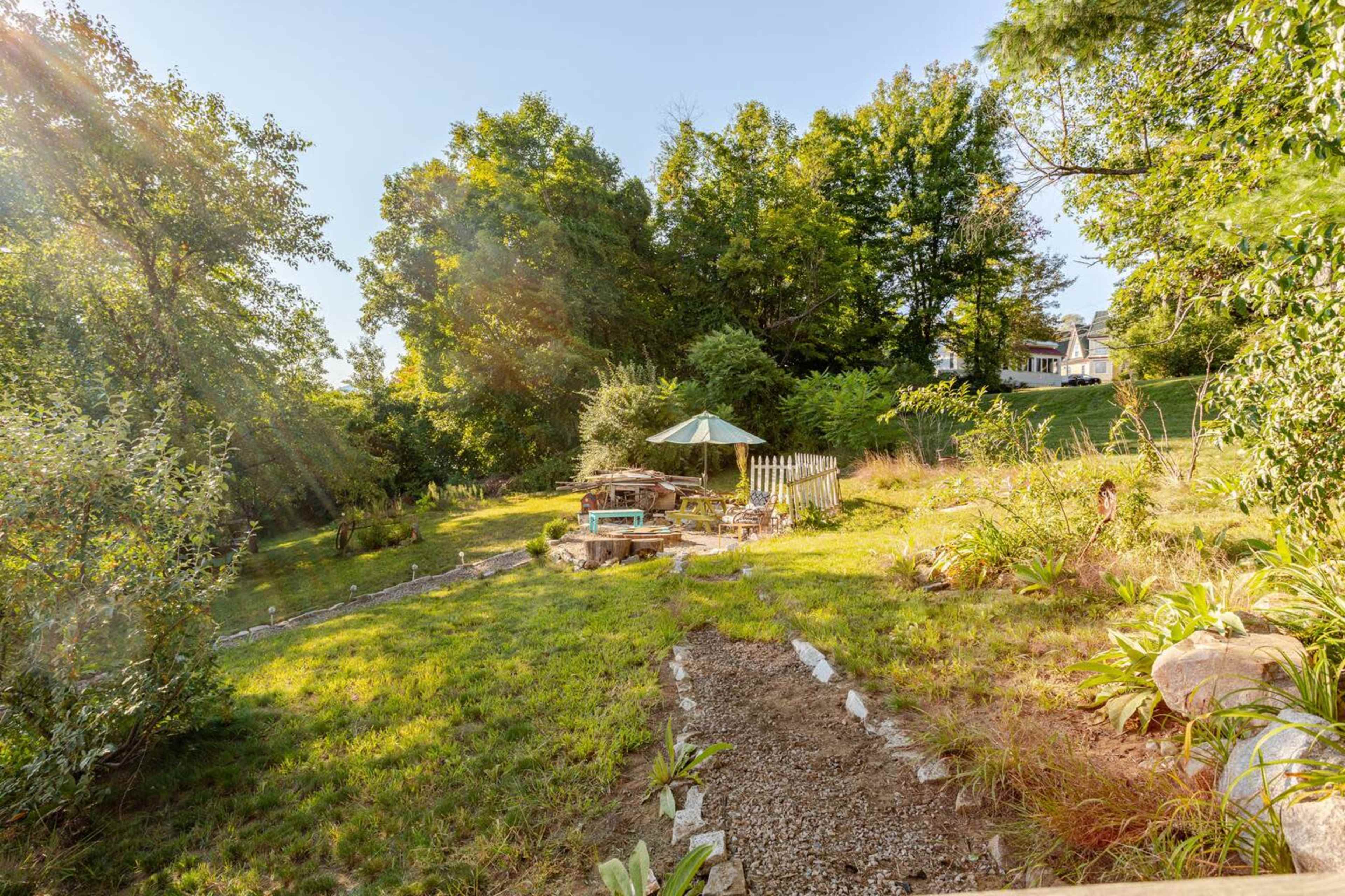 The image shows a landscaped backyard with a stone path leading to a patio area featuring a table and umbrella near a creek surrounded by trees.