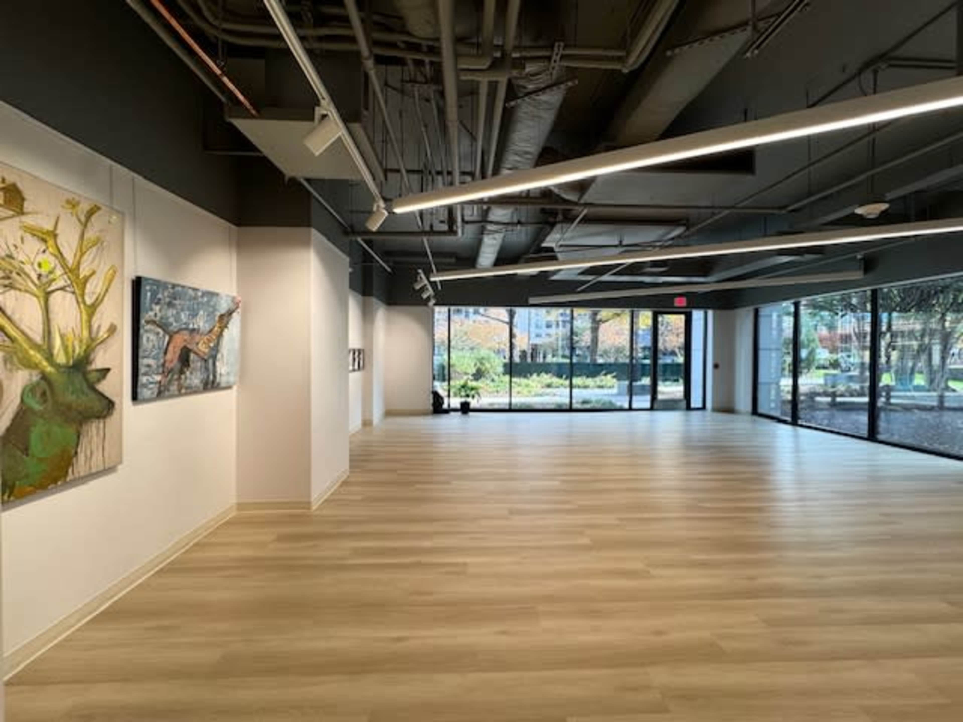 A spacious, well-lit gallery features wooden flooring, large windows, and artwork on the walls.
