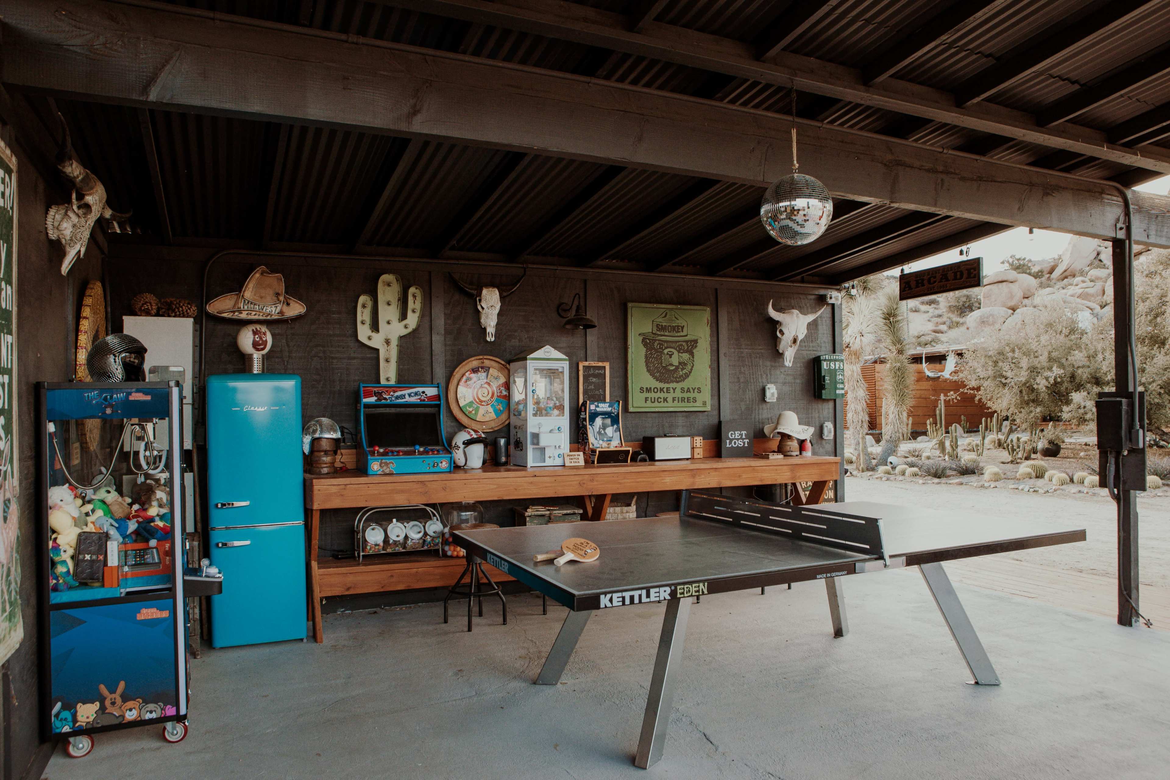 The image shows a covered outdoor area featuring a ping pong table, a vintage arcade machine, a blue refrigerator, and various decorative items, including cacti and animal skulls.