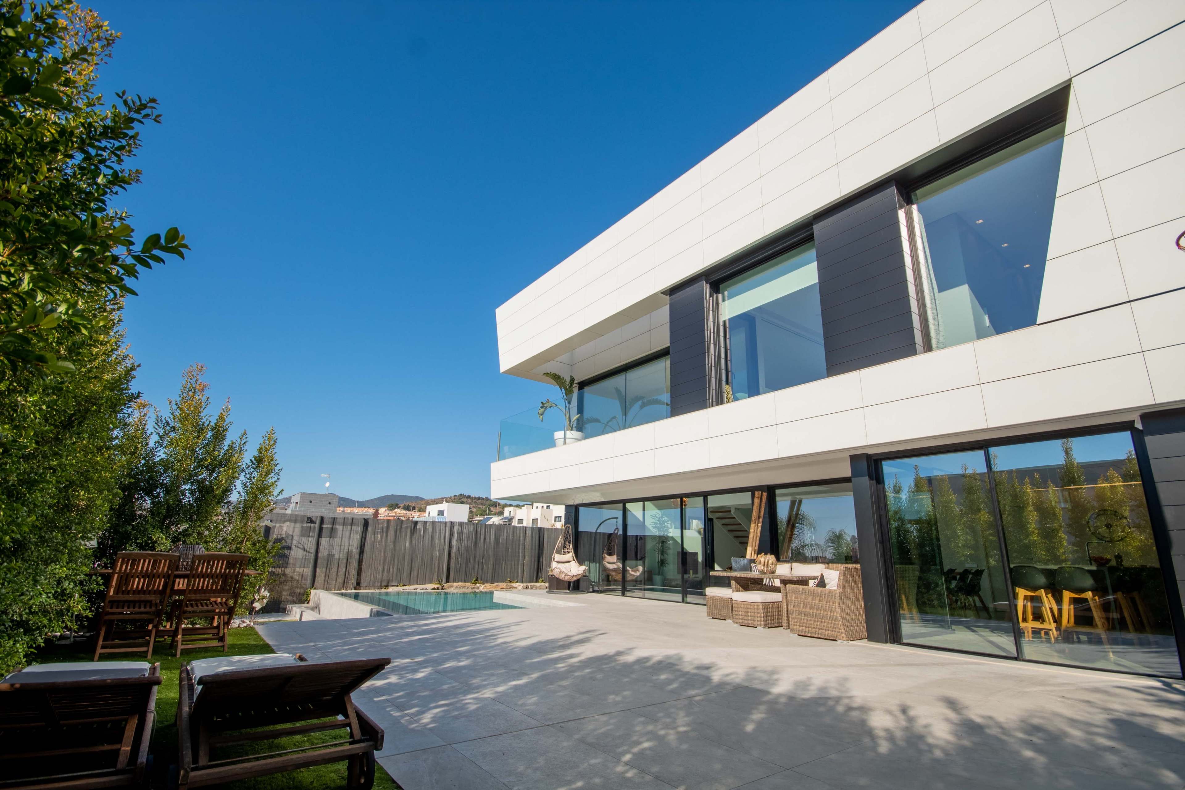 A modern two-story house with large glass windows, a swimming pool, and a spacious outdoor patio area surrounded by greenery.