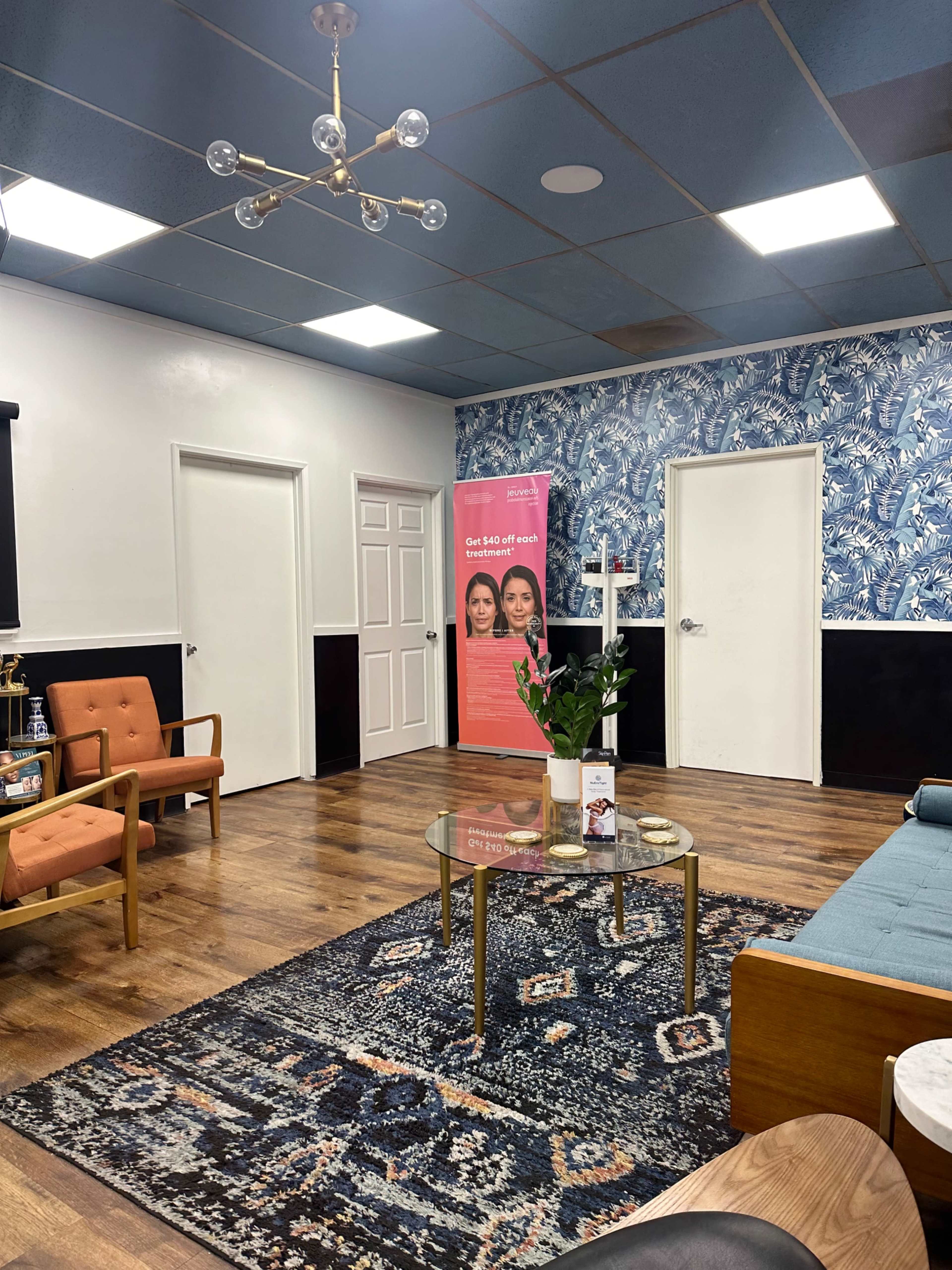A waiting room with blue and white patterned wallpaper, wooden flooring, a coffee table, and orange armchairs, featuring a promotional banner on the side.