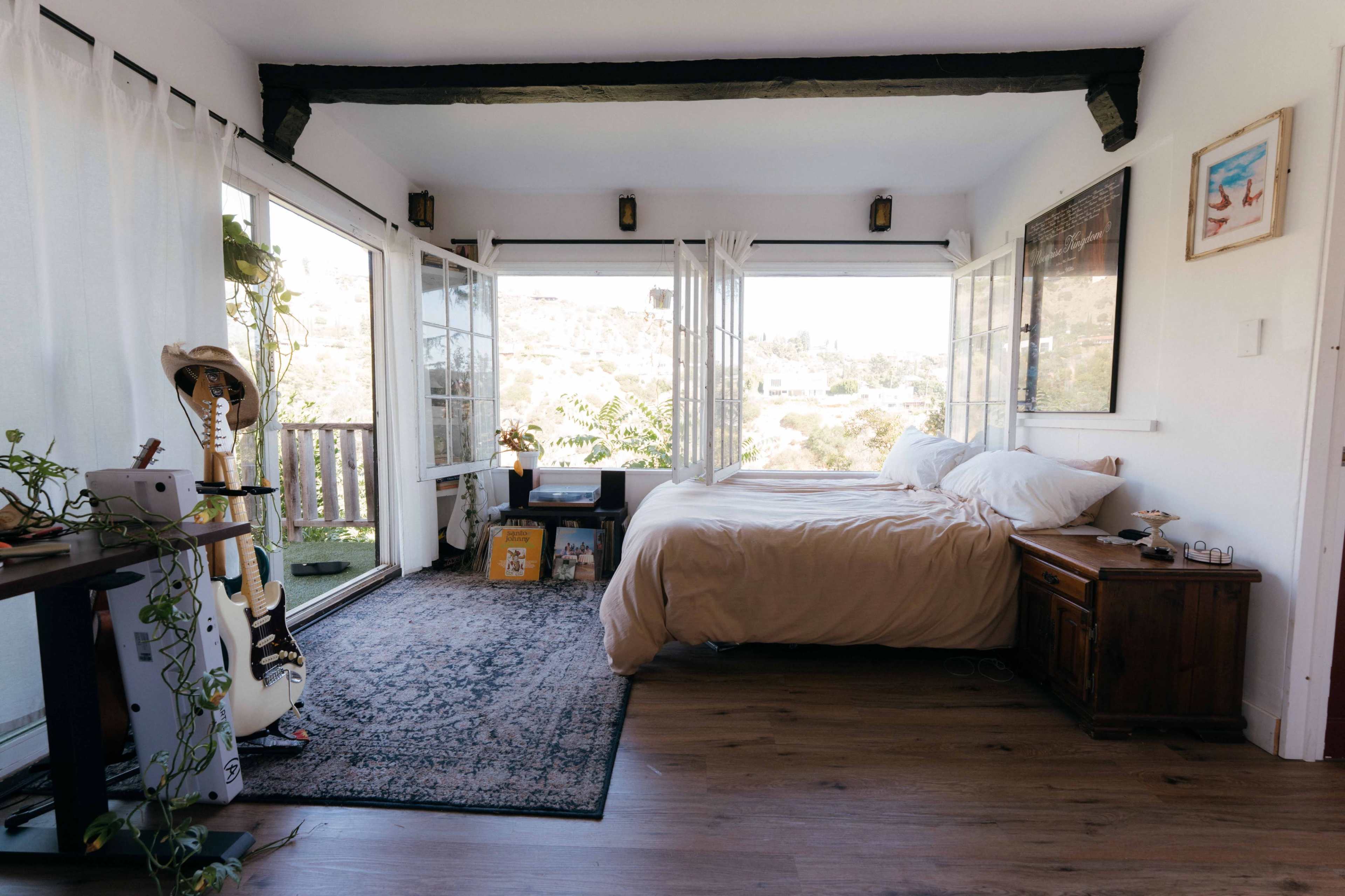 A cozy bedroom with large windows overlooking a hillside, featuring a bed, wooden furniture, and a guitar stand.