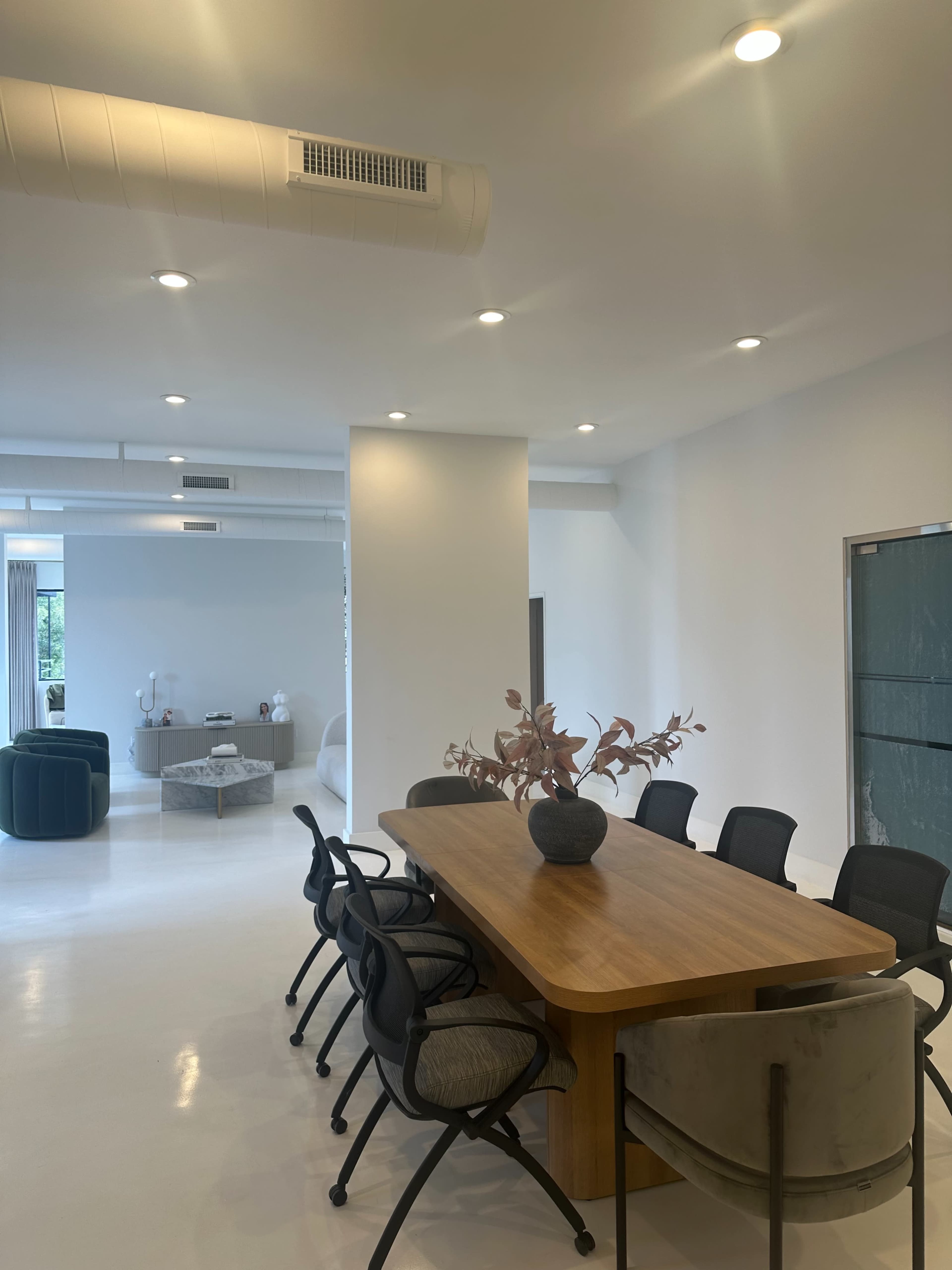 The image shows a modern dining area with a wooden table surrounded by black and gray chairs, brightened by overhead lights and minimalist decor.