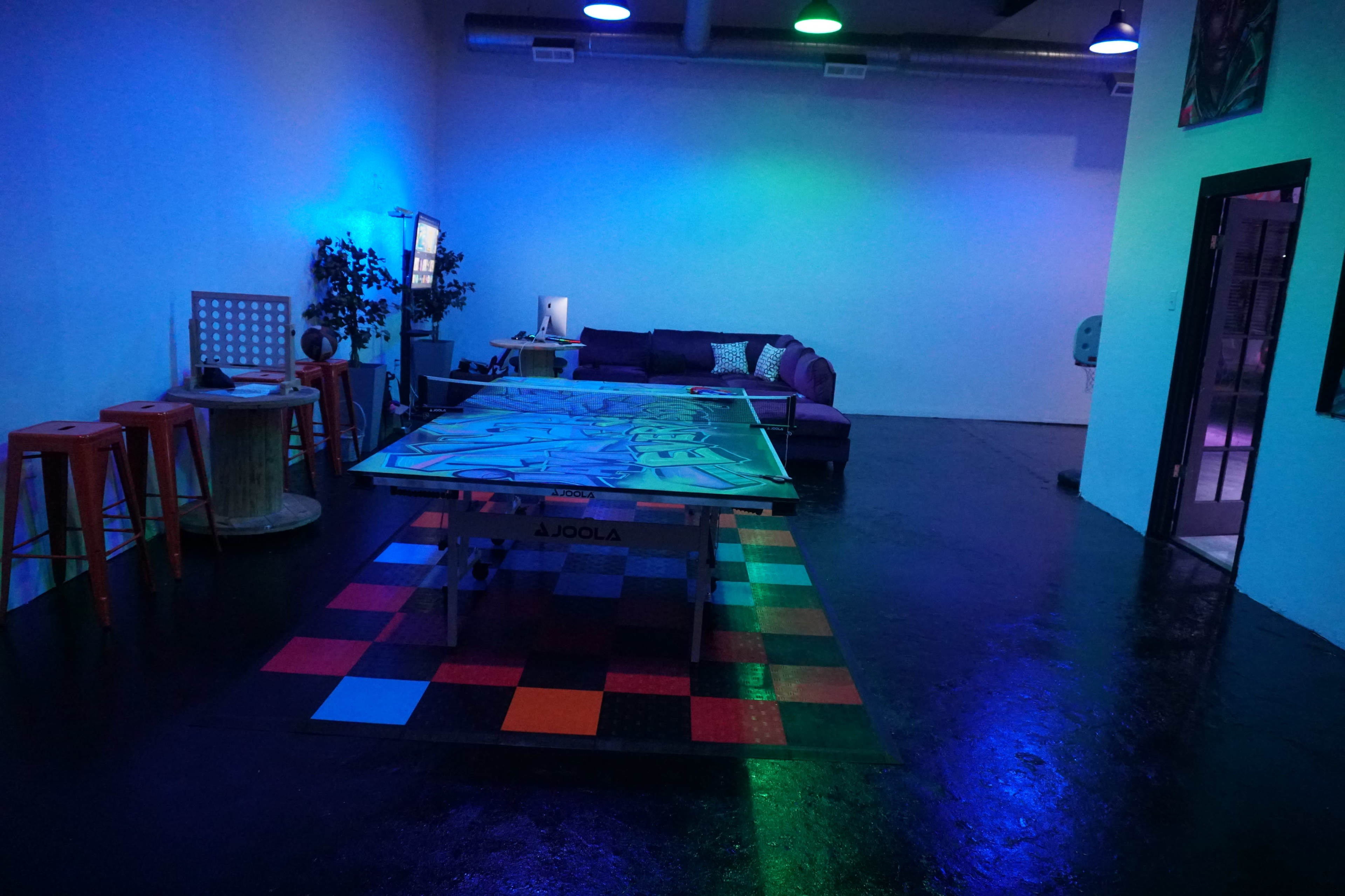 A dimly lit room featuring a ping pong table, colorful flooring, a couch, and various seating arrangements.