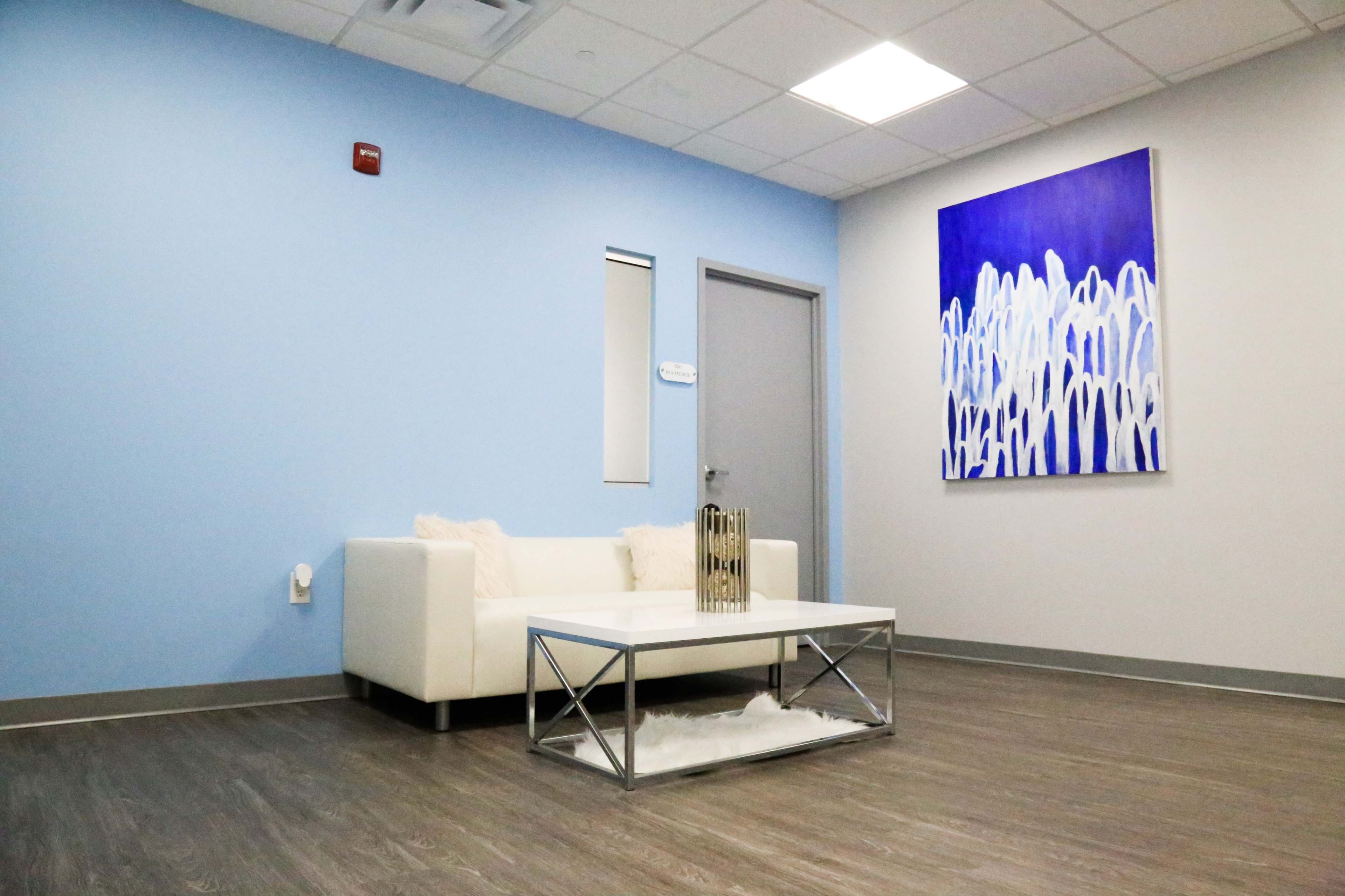 A modern waiting area featuring a white couch, a glass coffee table, and a blue wall with an abstract artwork.