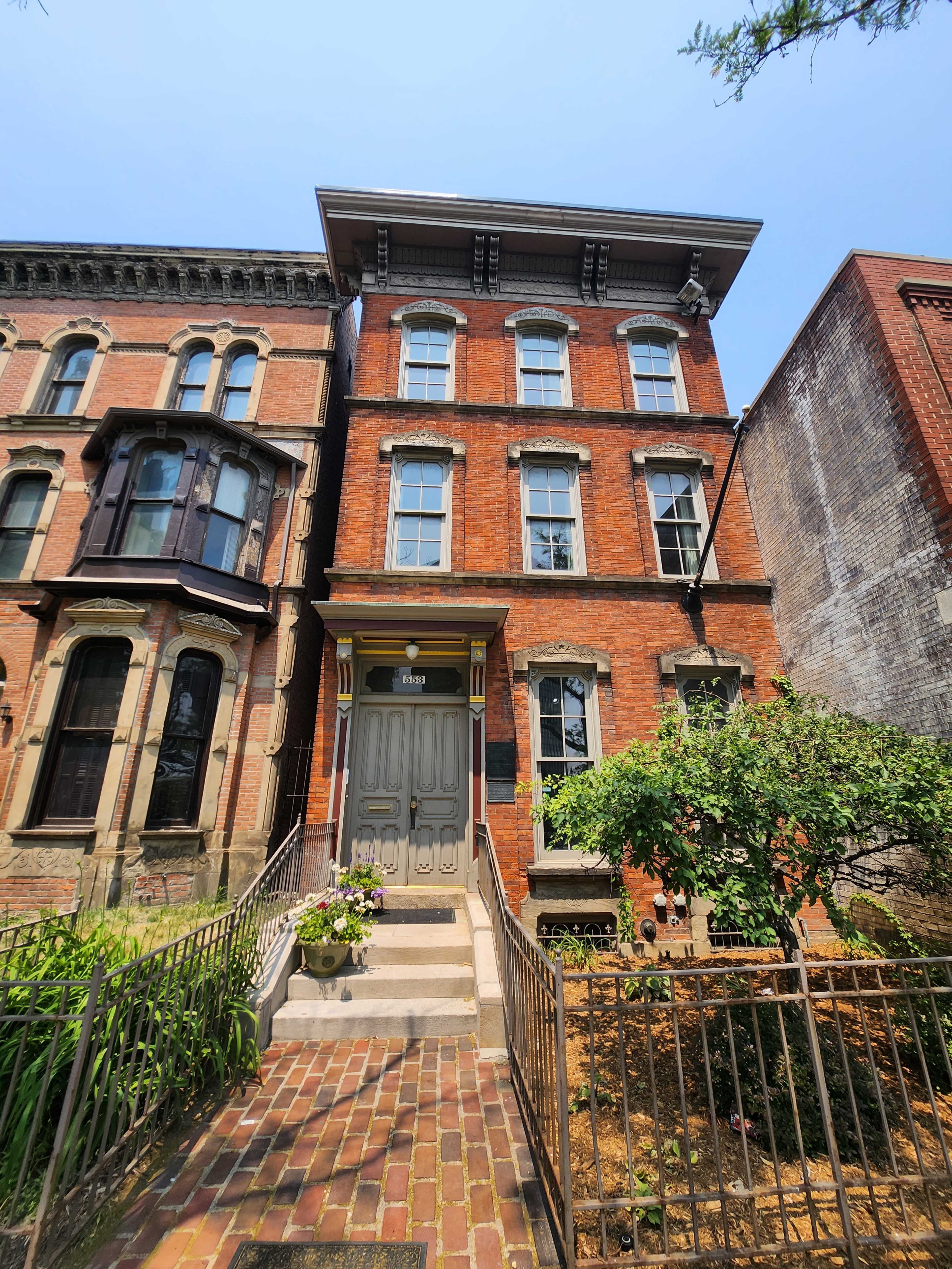 A three-story brick townhouse with large windows and a front staircase, flanked by a garden and neighboring buildings.