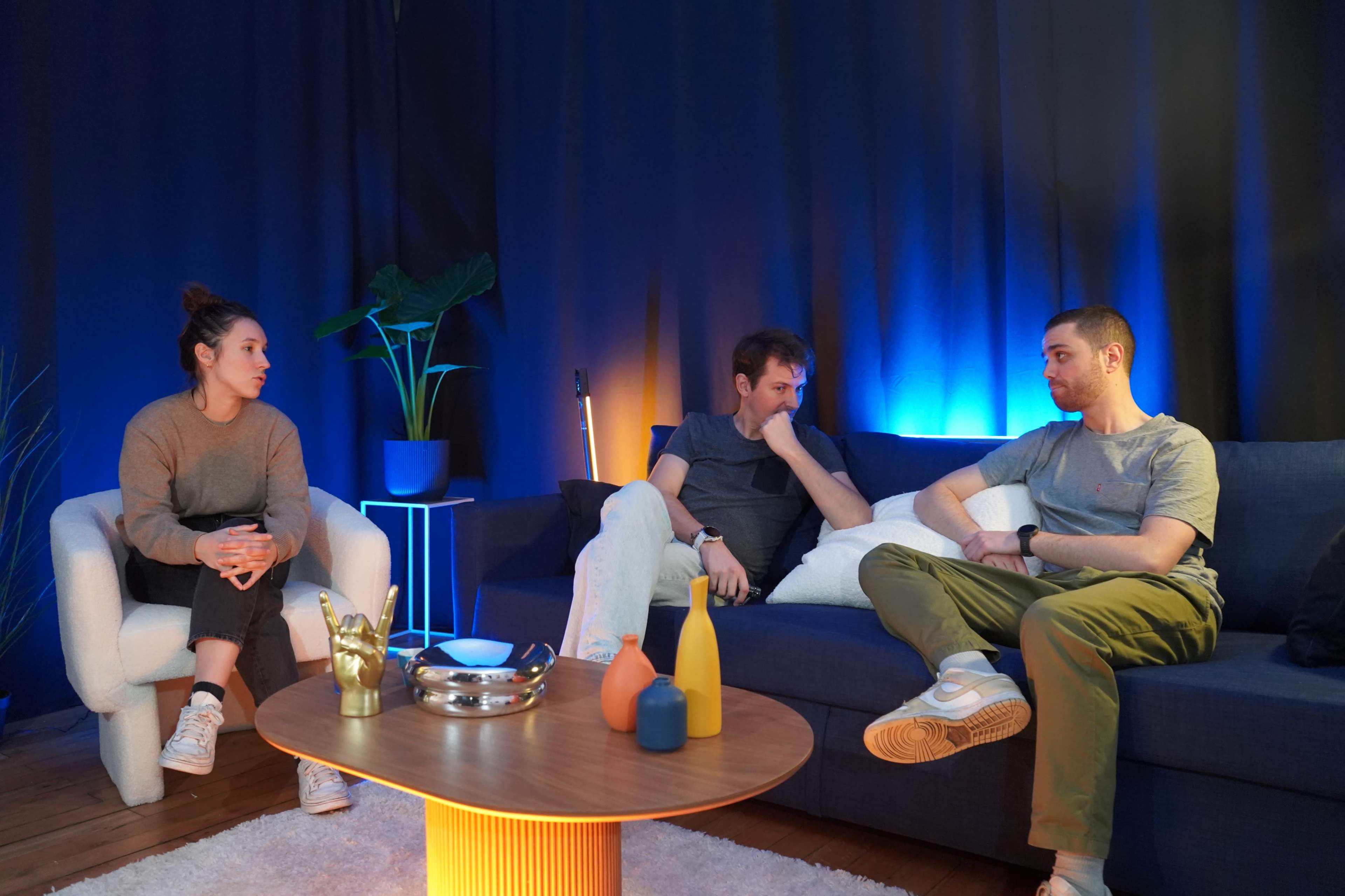 Three people sit on a sofa in a dimly lit room with blue and orange lighting, engaged in conversation.