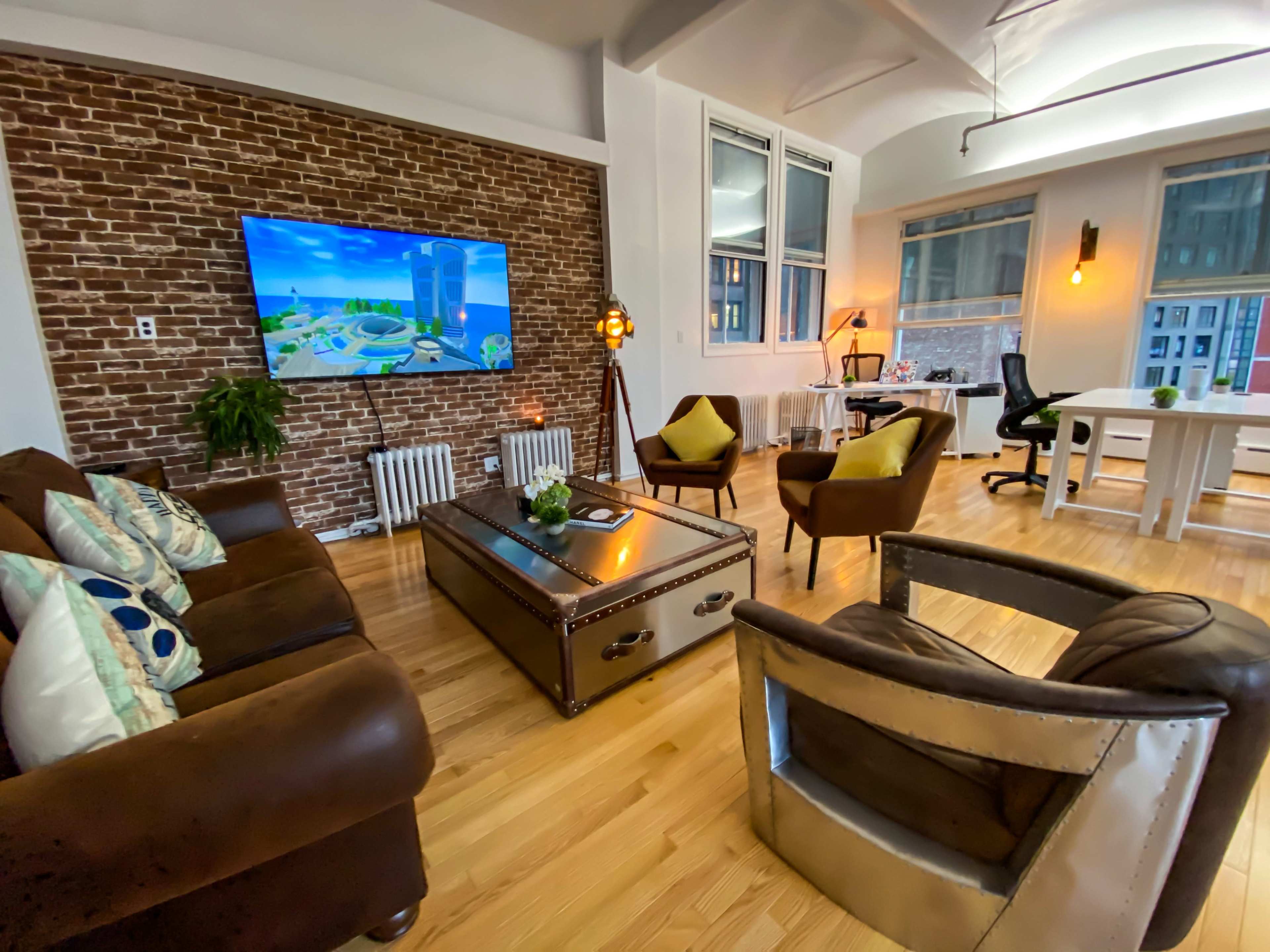 A modern living room with exposed brick walls, a large television, comfortable seating, and a workspace featuring a desk and office chair.