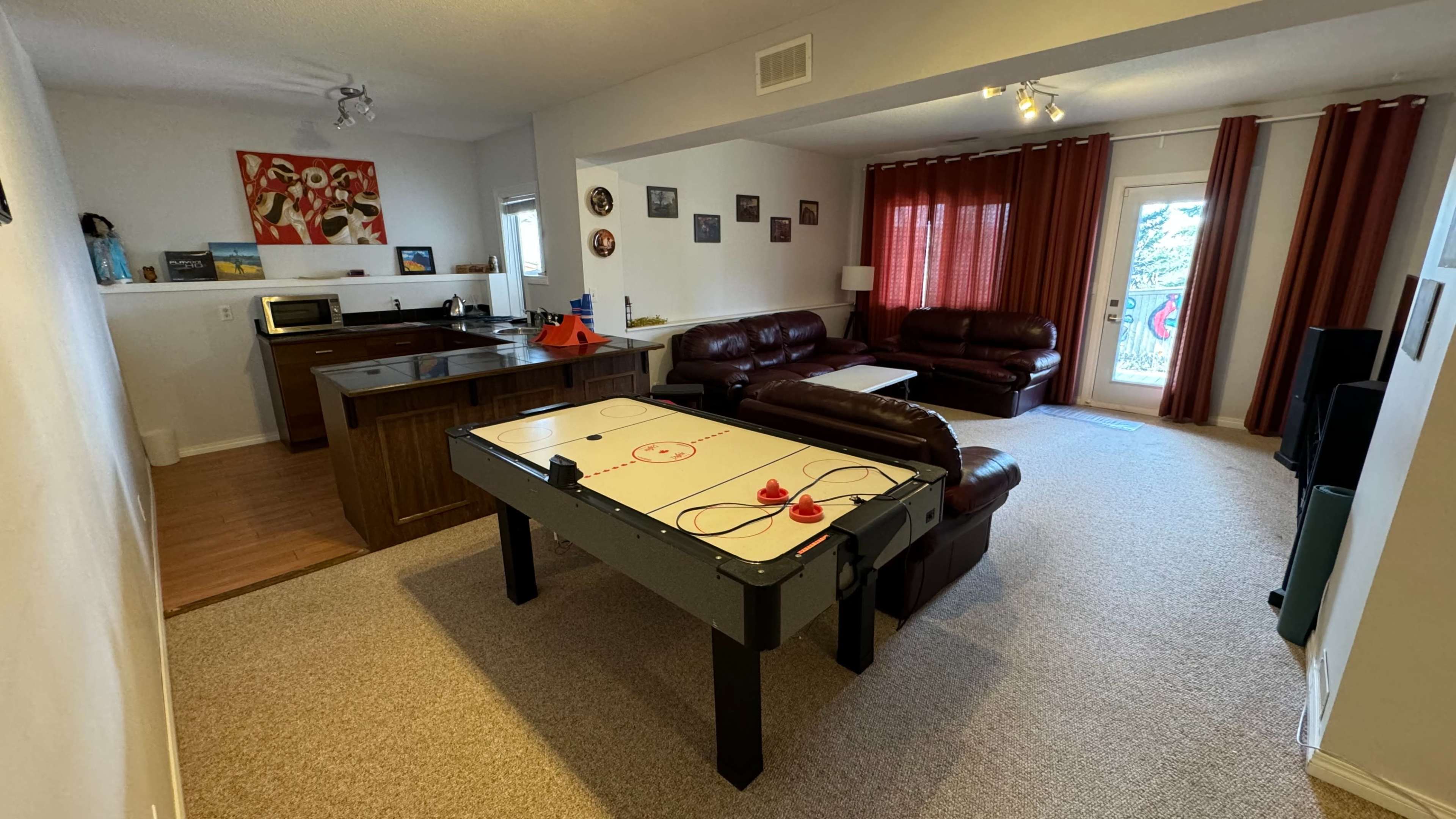 A cozy living room featuring an air hockey table, a dark leather sofa set, and a kitchen area in the background.