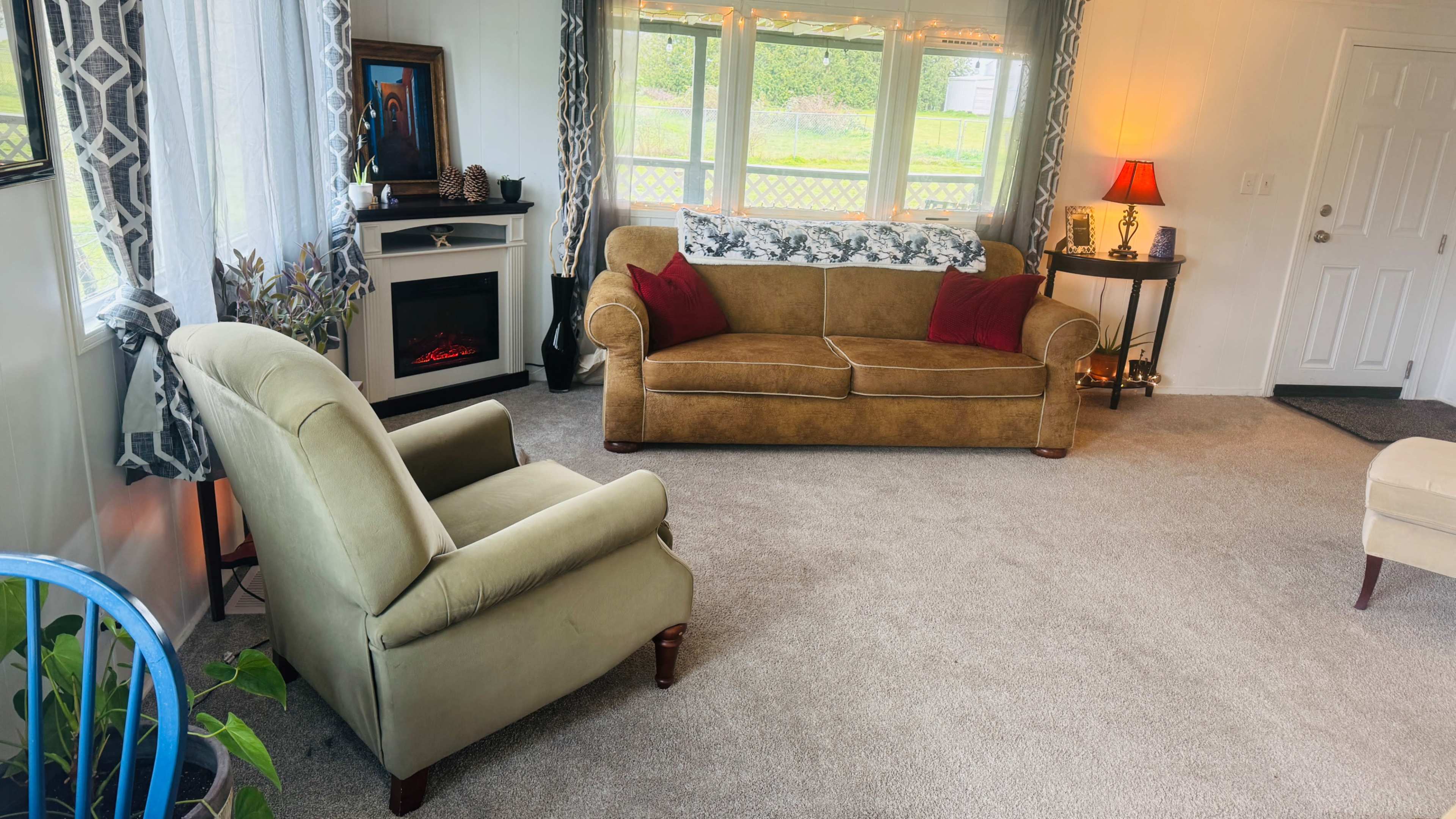 The living room features a brown sofa with red cushions and a green armchair, complemented by a small table with a lamp and a fireplace, all set against a backdrop of large windows.