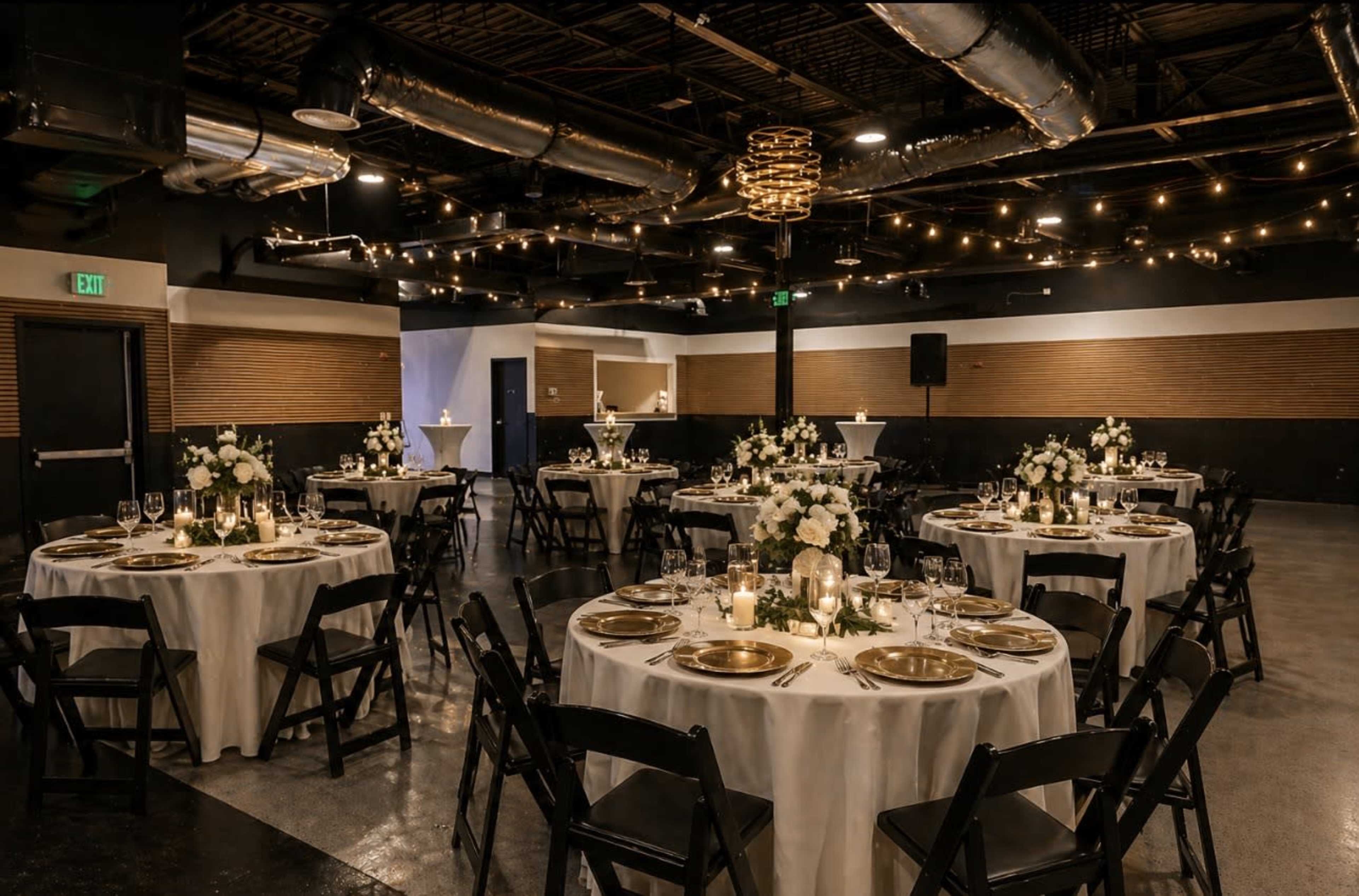 The image shows a decorated event space with round tables covered in white tablecloths, each set with gold plates and floral centerpieces, under warm ambient lighting.