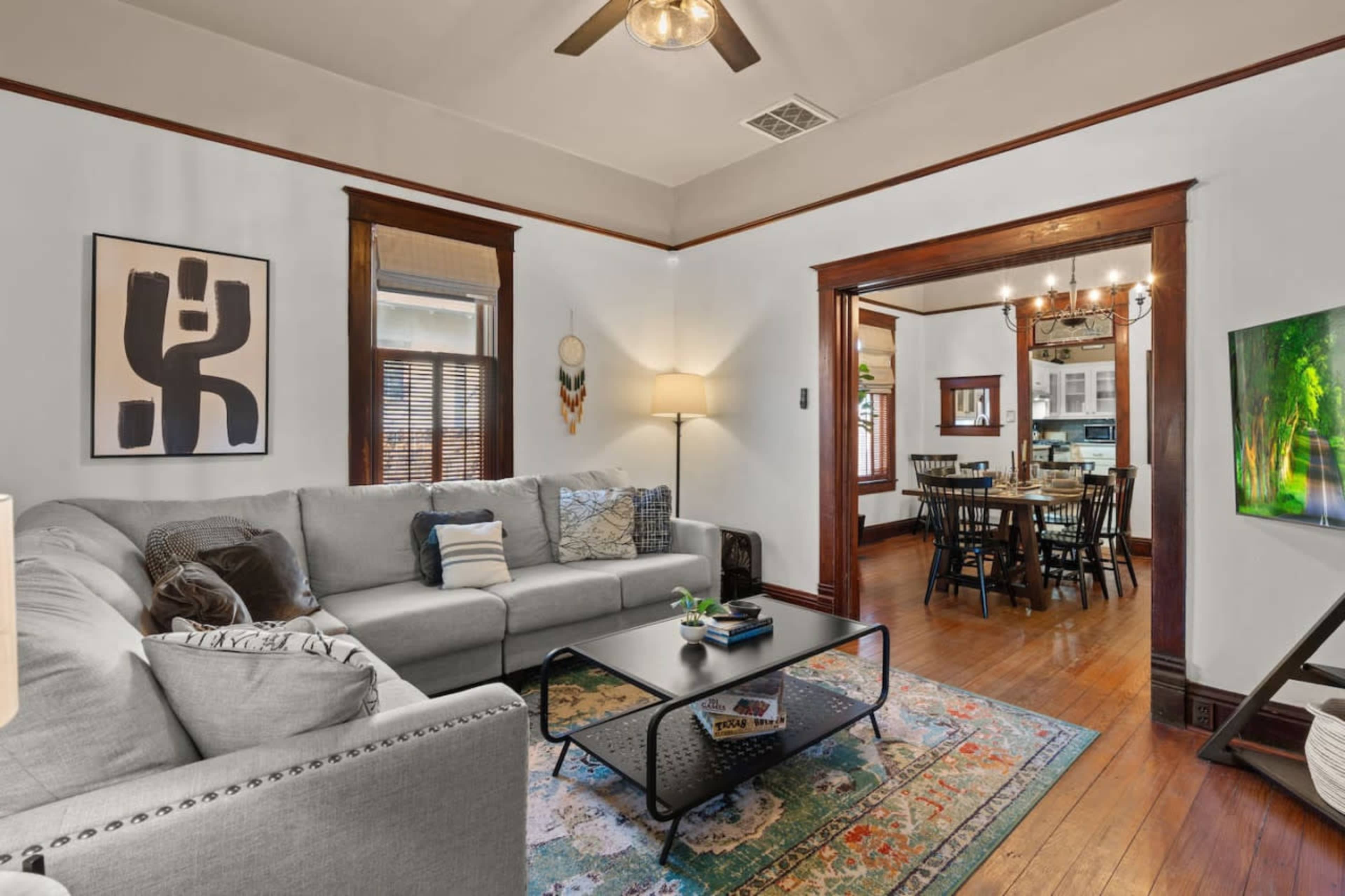A living room with a gray sectional sofa, a black coffee table, and a decorative area rug, leading to a dining area visible through an opening.