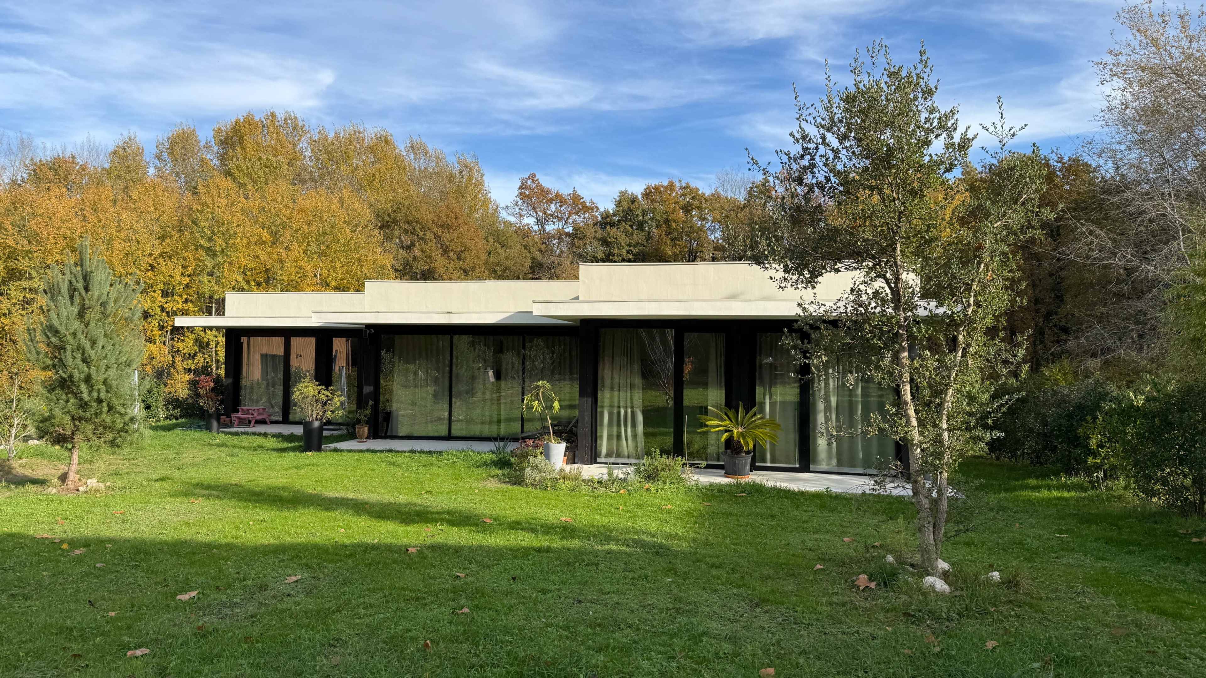 A modern, single-story house with large windows sits amidst a backdrop of colorful autumn trees and a green lawn.