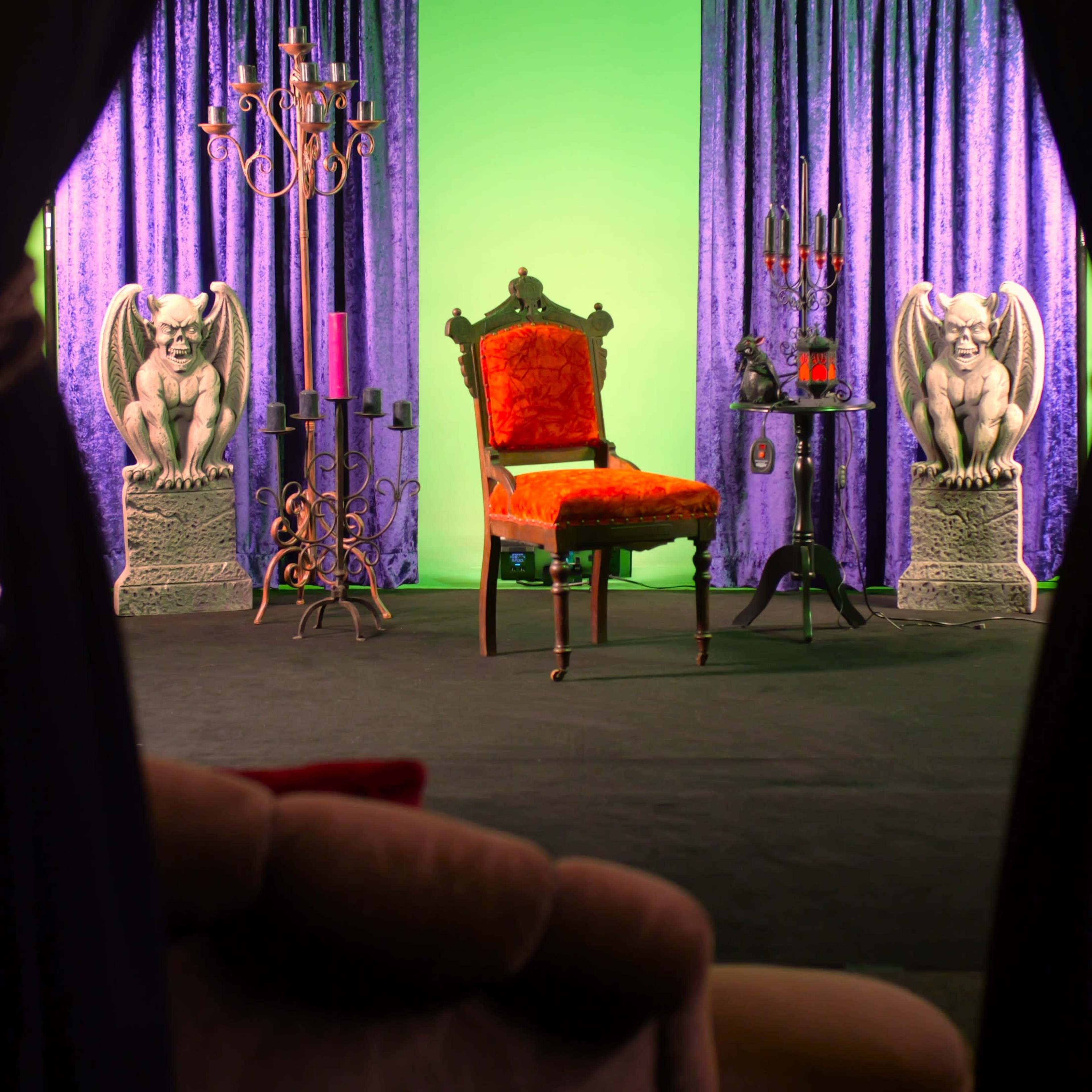 The scene features an ornate red chair positioned on a stage with purple curtains, flanked by two gargoyle statues and a candelabrum, set against a green screen backdrop.