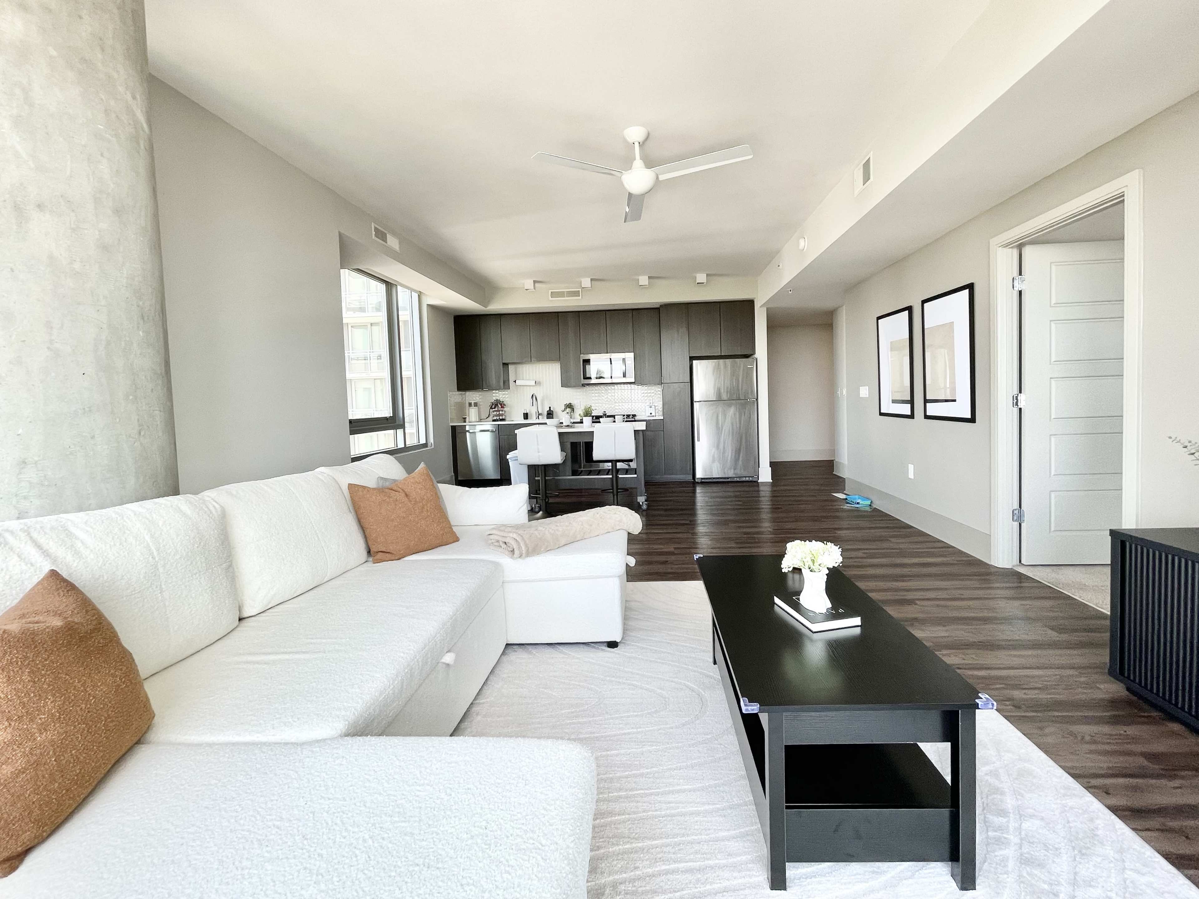 The living area features a white sectional sofa with a brown accent pillow, a dark coffee table, and a kitchen area visible in the background.