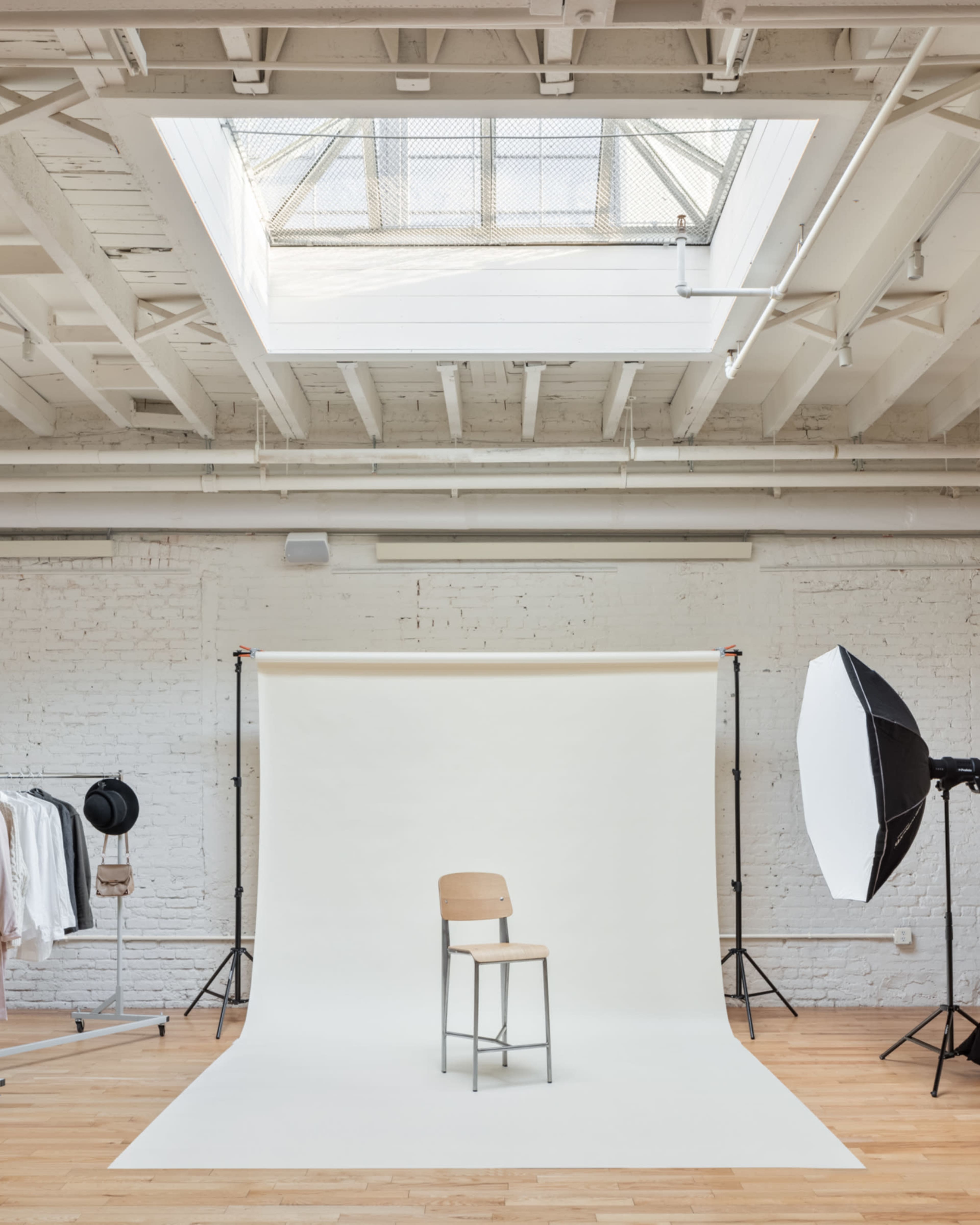 A wooden chair is positioned in front of a white backdrop in a well-lit photography studio with a skylight and studio equipment.