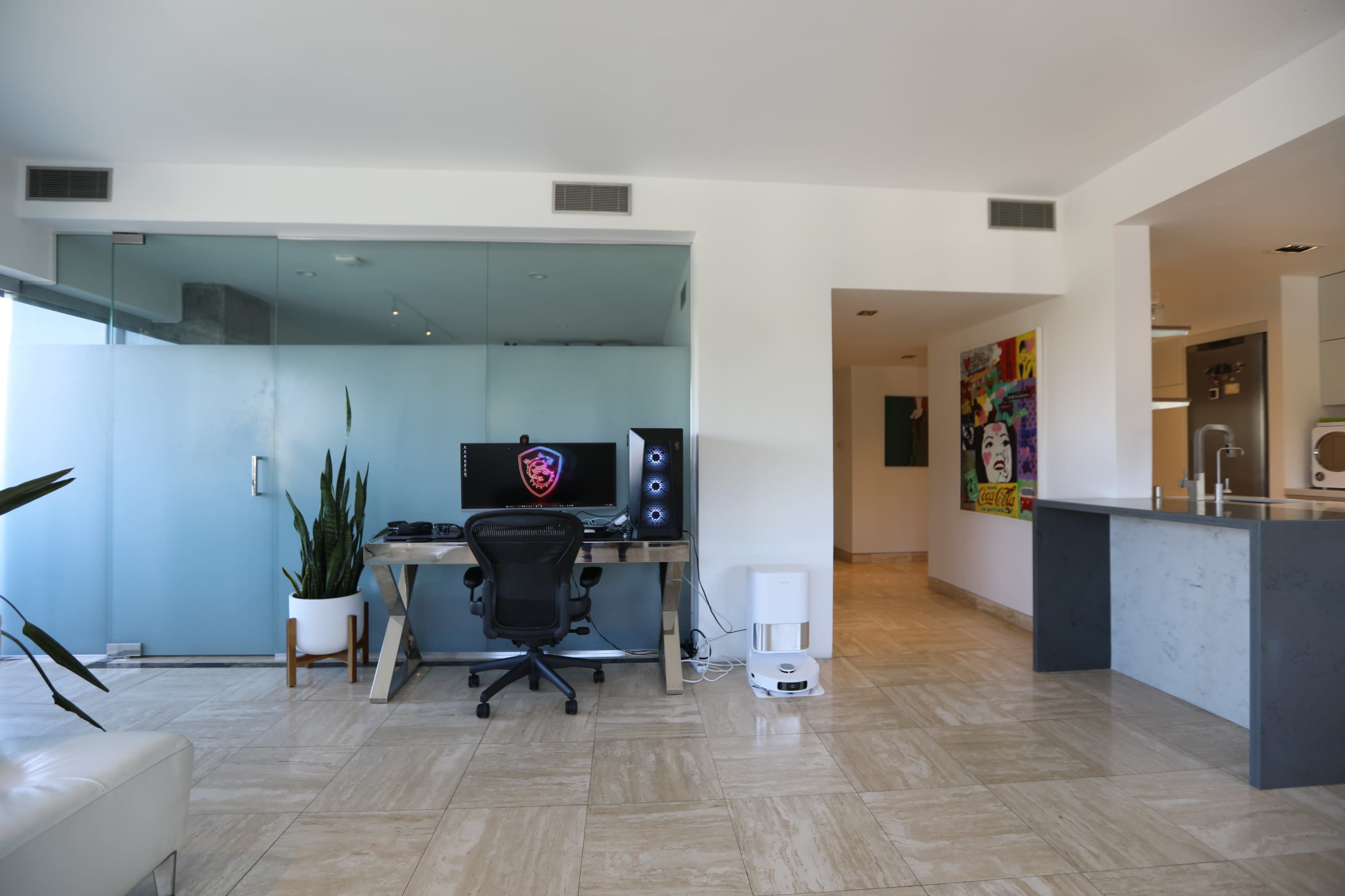 The image shows a modern apartment interior featuring a workspace with a computer setup, a plant, and an open layout connecting to a kitchen area.