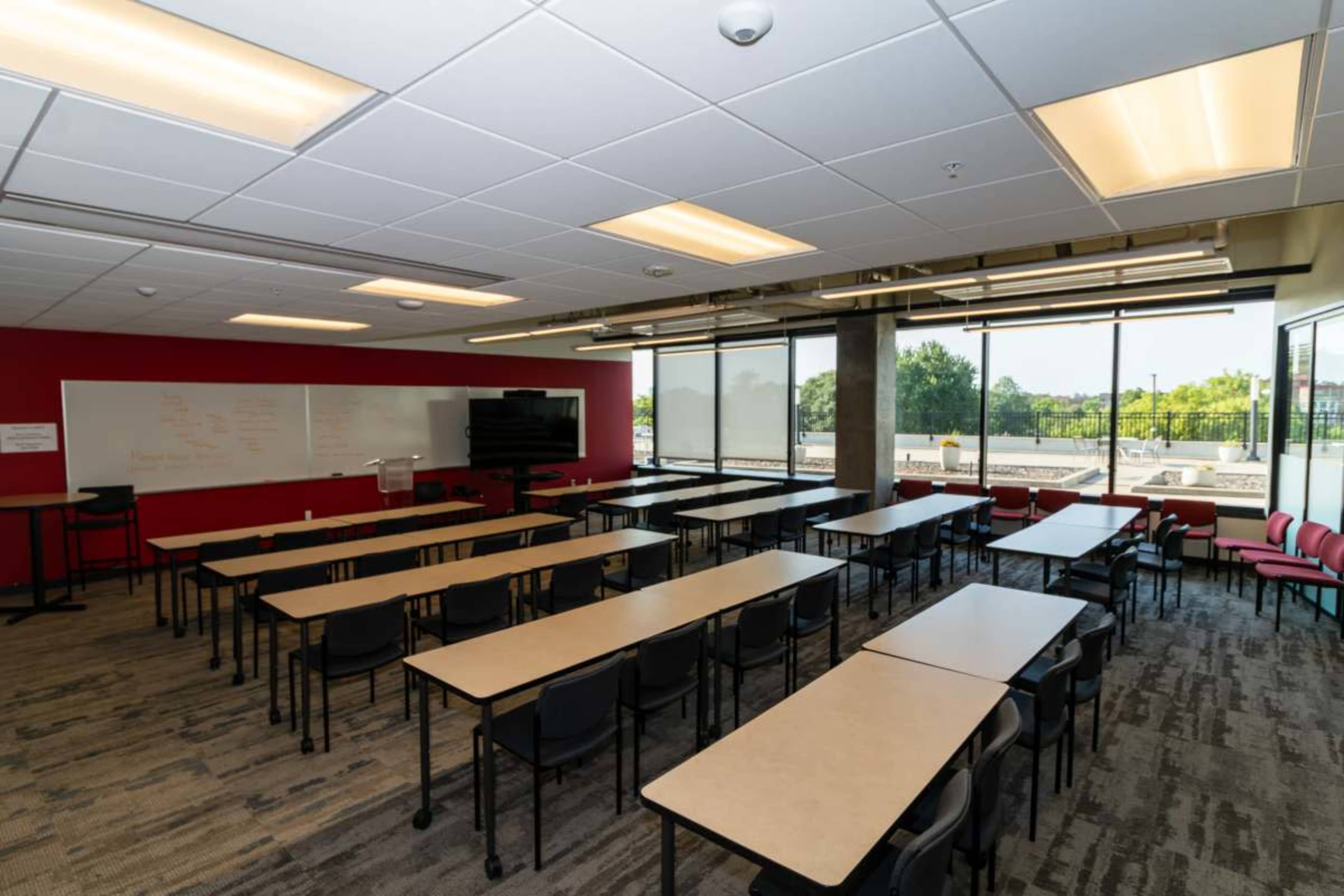 The image shows a modern classroom setup with rows of rectangular tables and chairs, large windows providing ample natural light, and a whiteboard on one wall.
