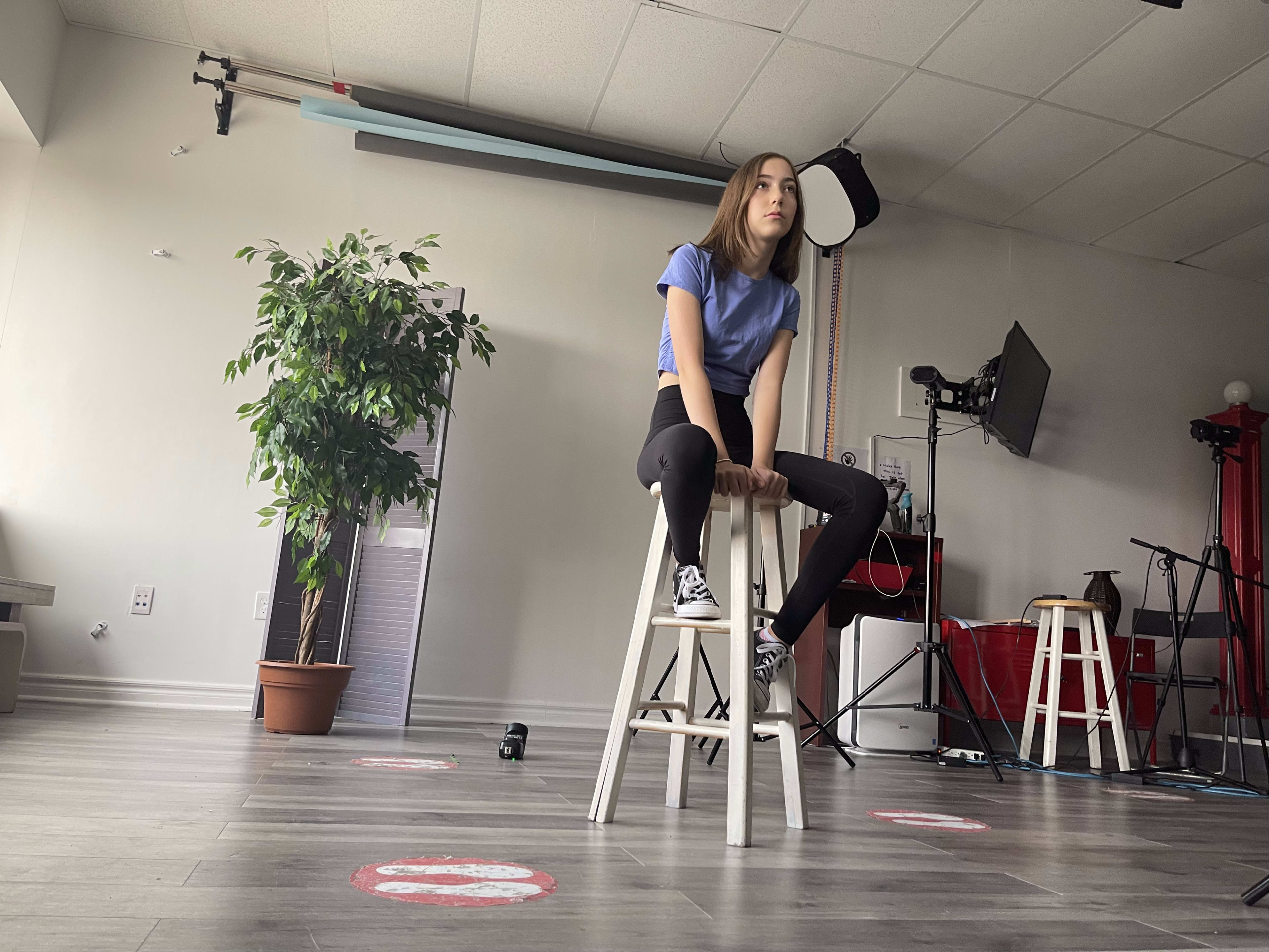 A young woman sits on a wooden stool in a studio with a plant and various equipment in the background.