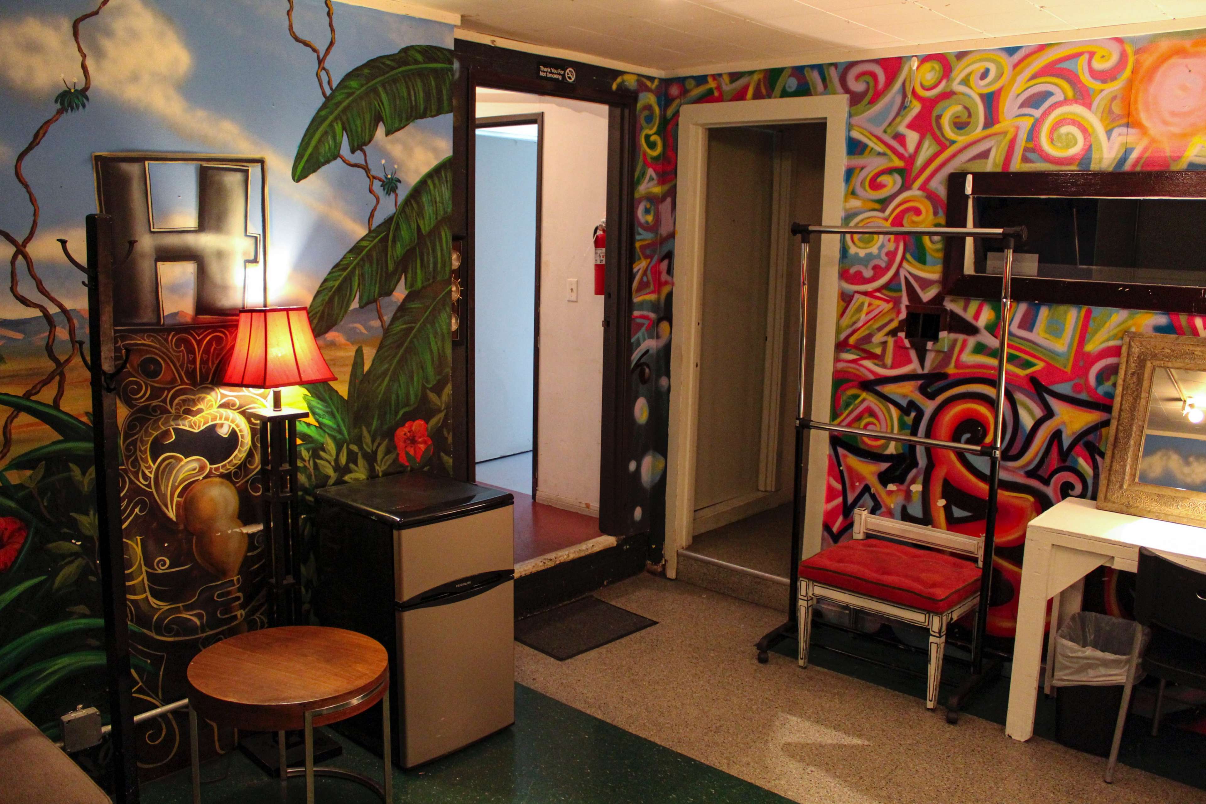 The room features colorful murals on the walls, a small table with a lamp, a mini fridge, and a folding chair set against a backdrop of various artistic designs.