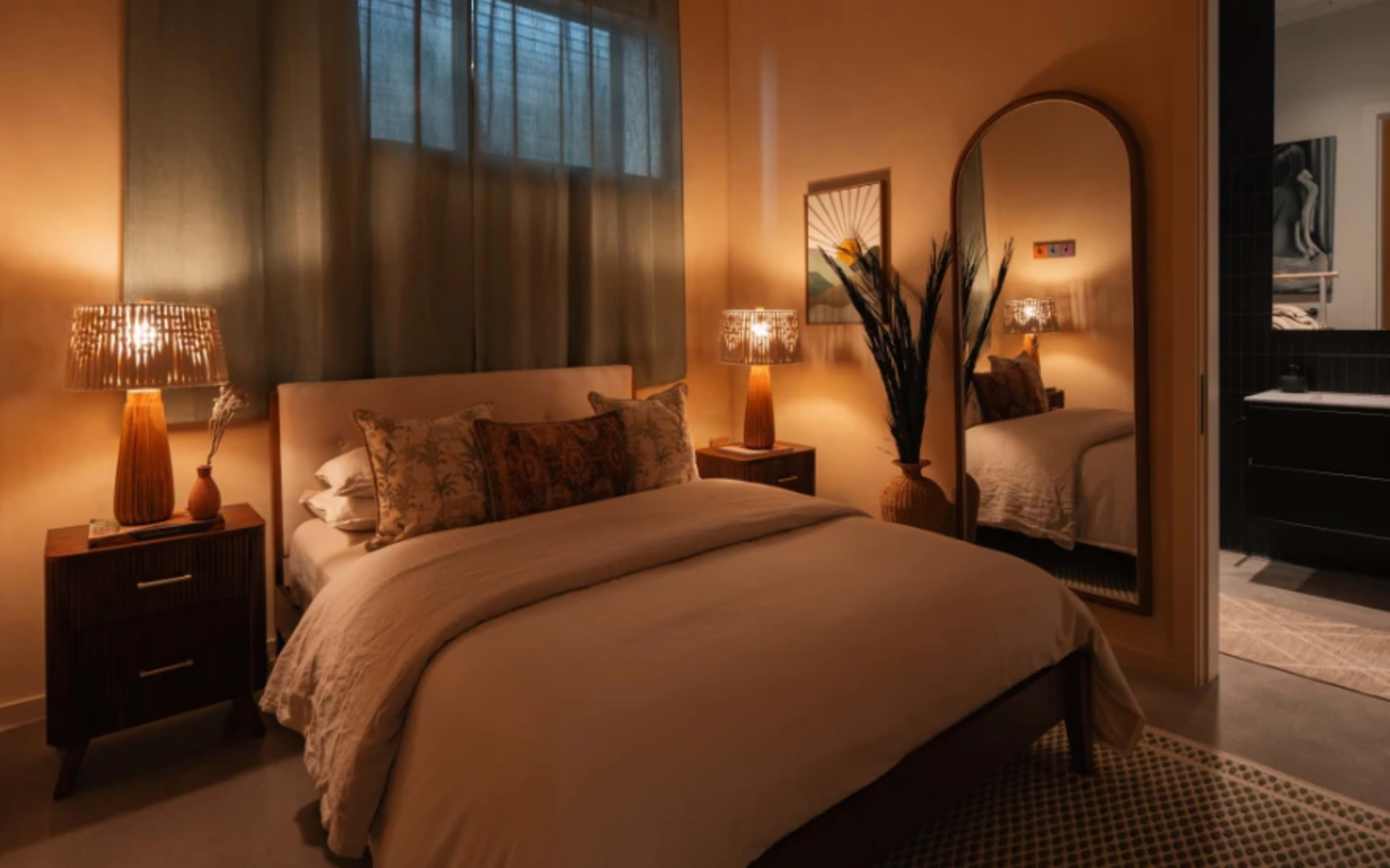 The image shows a warmly lit bedroom featuring a large bed with decorative pillows, two bedside lamps, and a mirror reflecting part of the room.