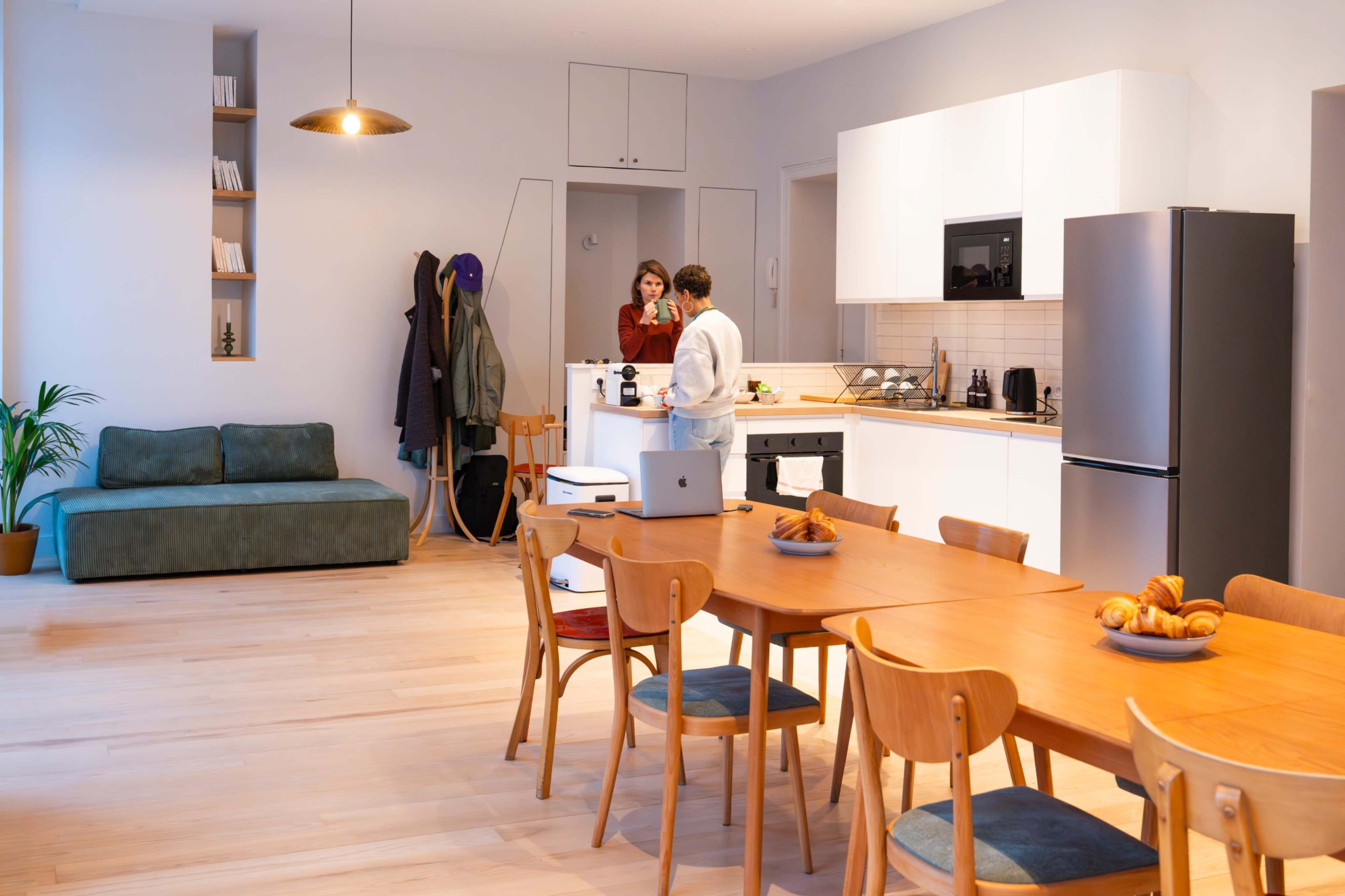 A modern kitchen and dining area features a wooden table surrounded by chairs, with a woman and man preparing food while a couch sits in the background.