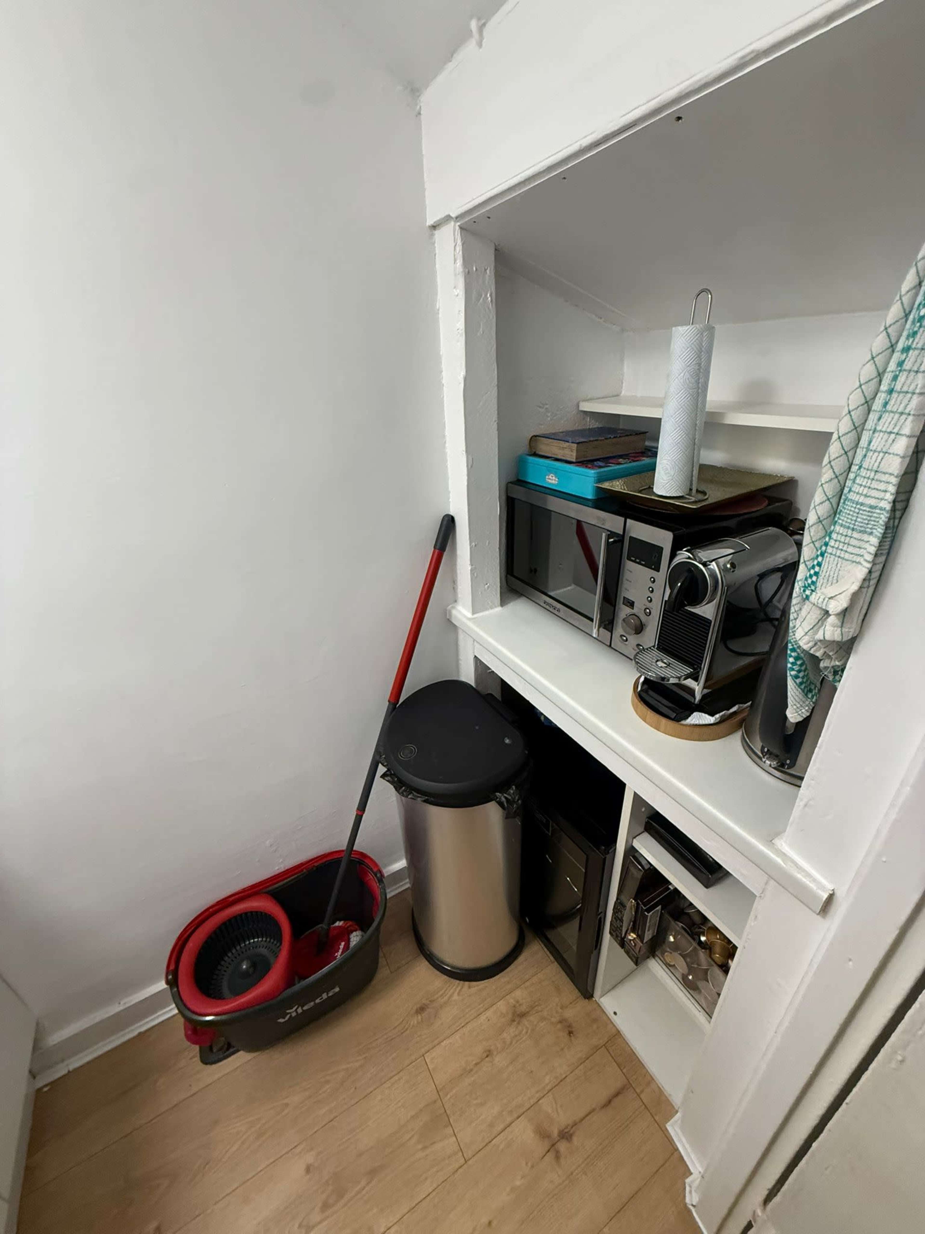 The image shows a small storage area featuring a vacuum cleaner, trash can, microwave, and several shelves with various kitchen items.