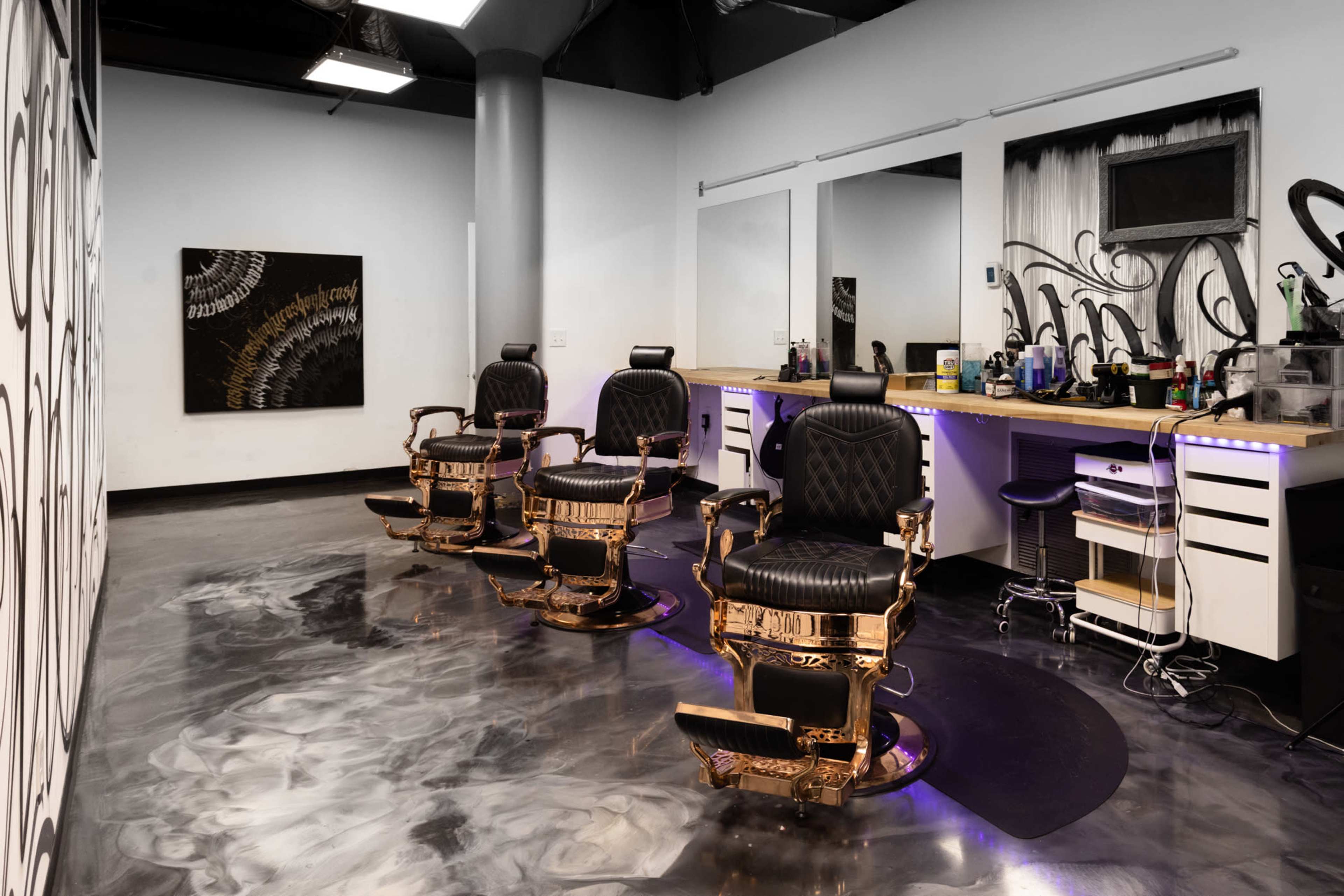 The image shows a modern barbershop interior featuring three golden barber chairs, a long countertop with various grooming products, and a decorative wall.