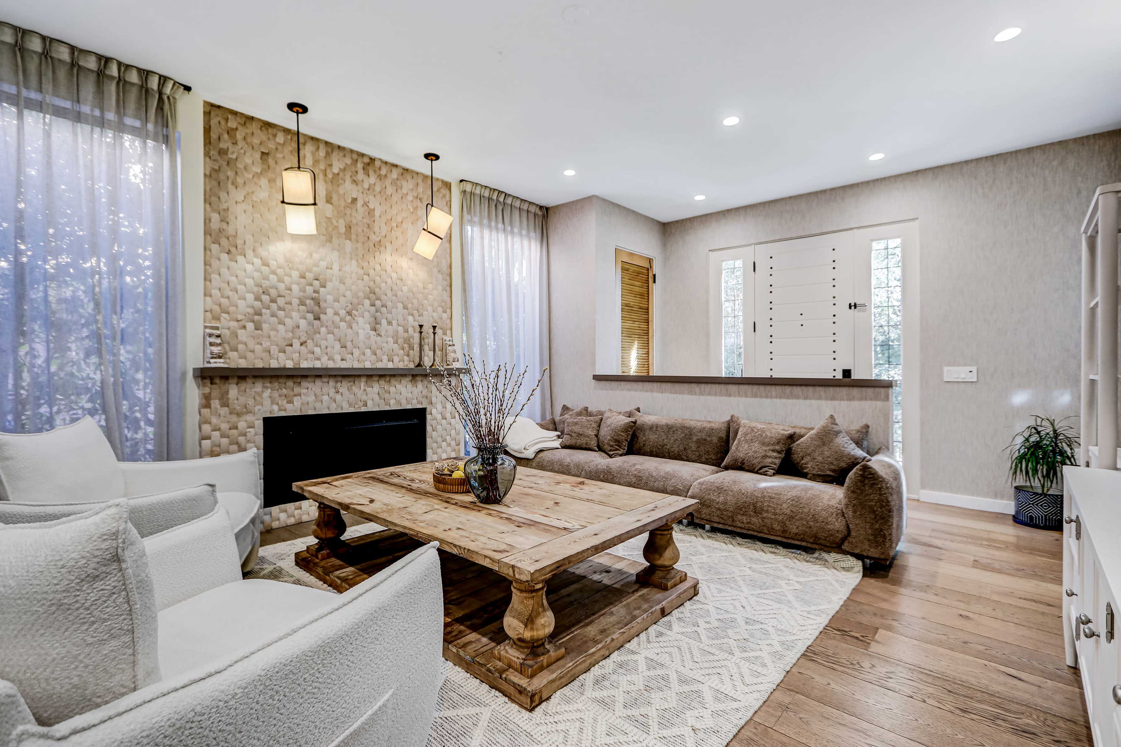 The living room features a large, rustic wooden coffee table surrounded by a sectional couch, with soft lighting and large windows allowing natural light to filter in.