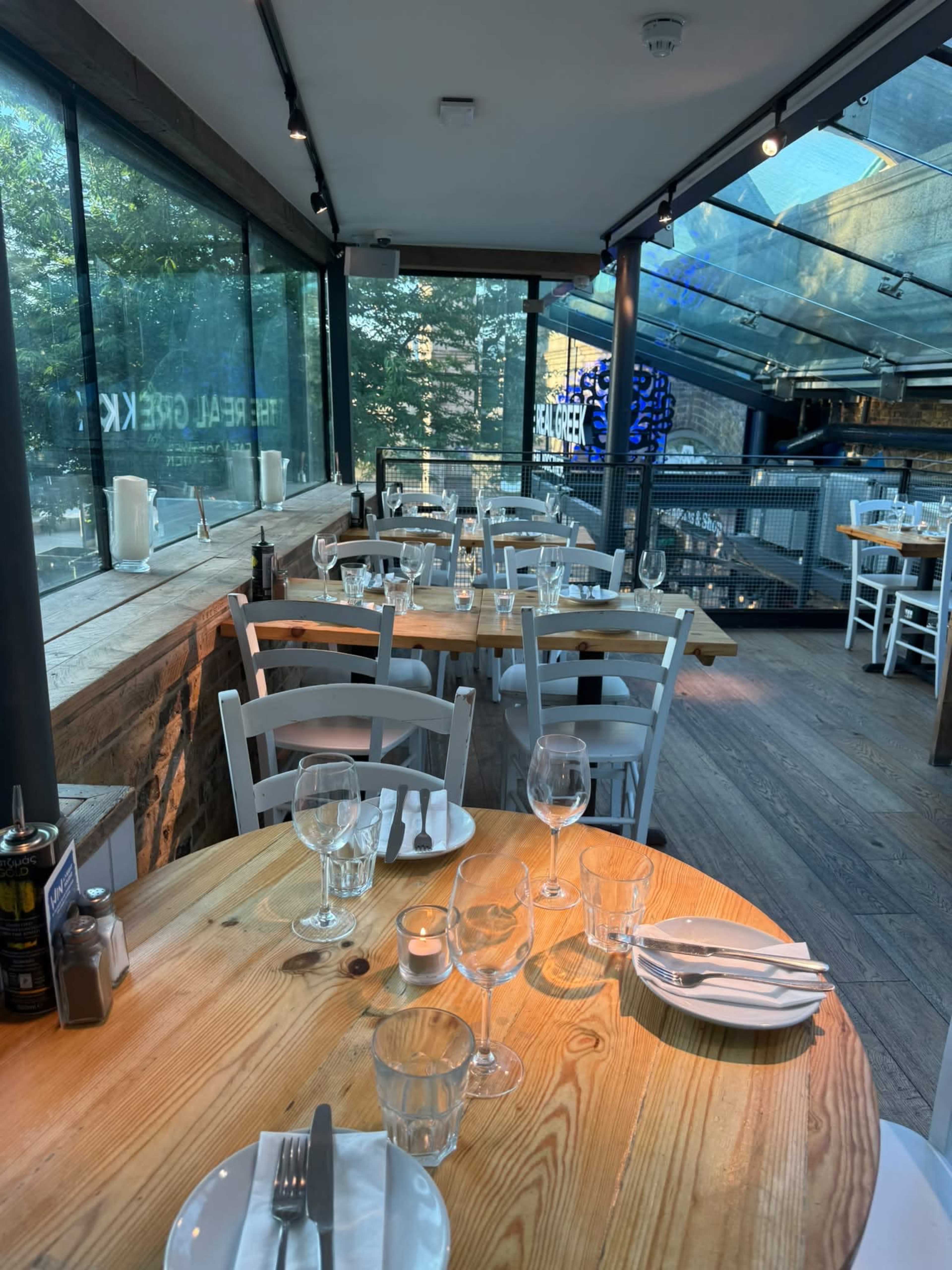 The image shows a dining area in a restaurant, featuring wooden tables, glass walls, and a contemporary design with tables set for guests.