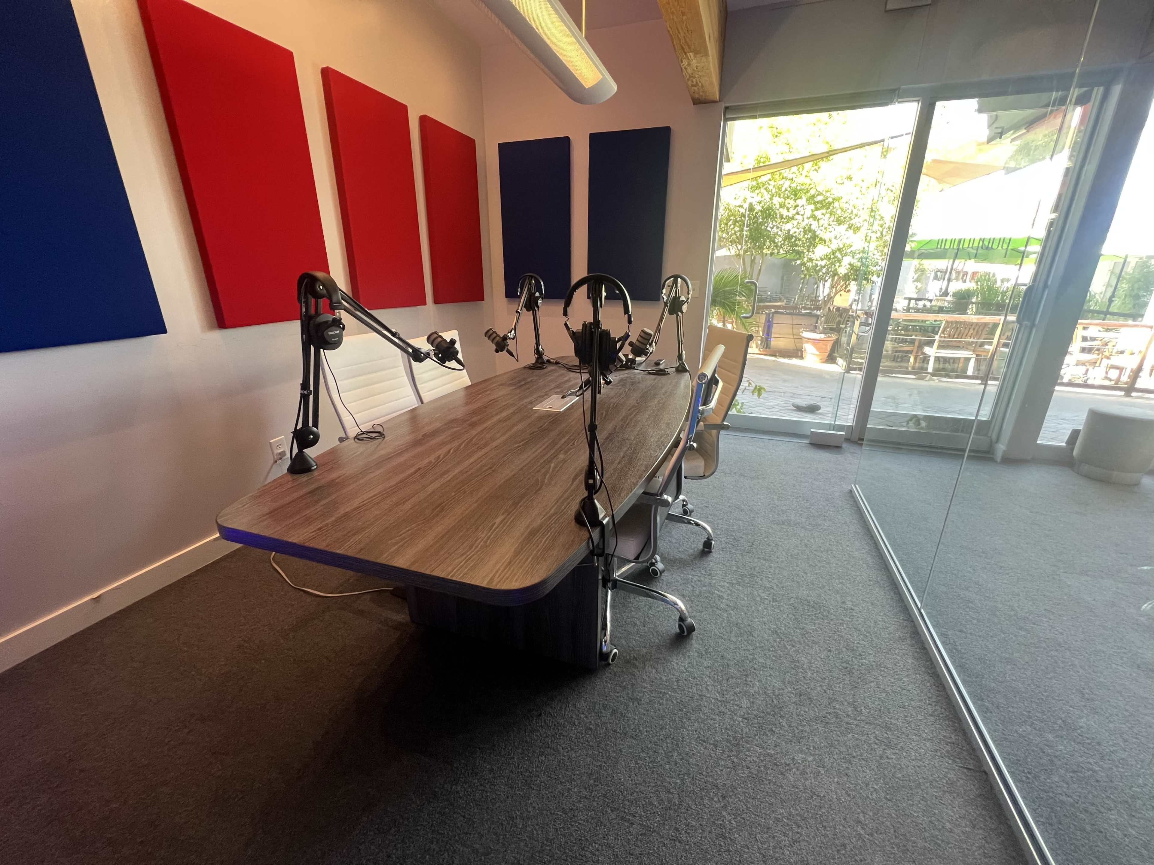 A modern recording studio features a large table with microphones and headphones, accented by colorful sound panels and a view of an outdoor area.