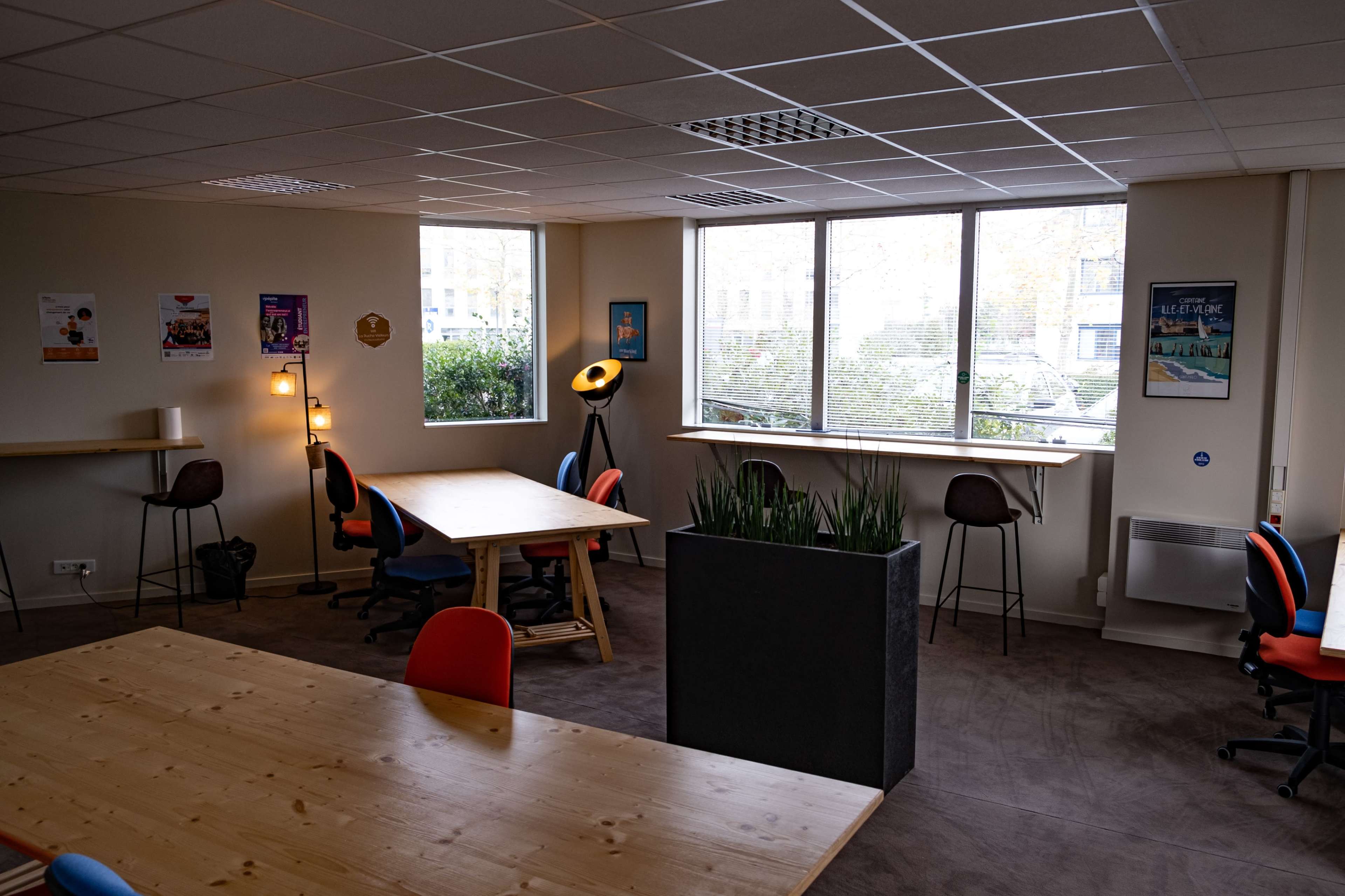 The image shows an open office space with several tables, chairs in various colors, and large windows allowing natural light to enter.