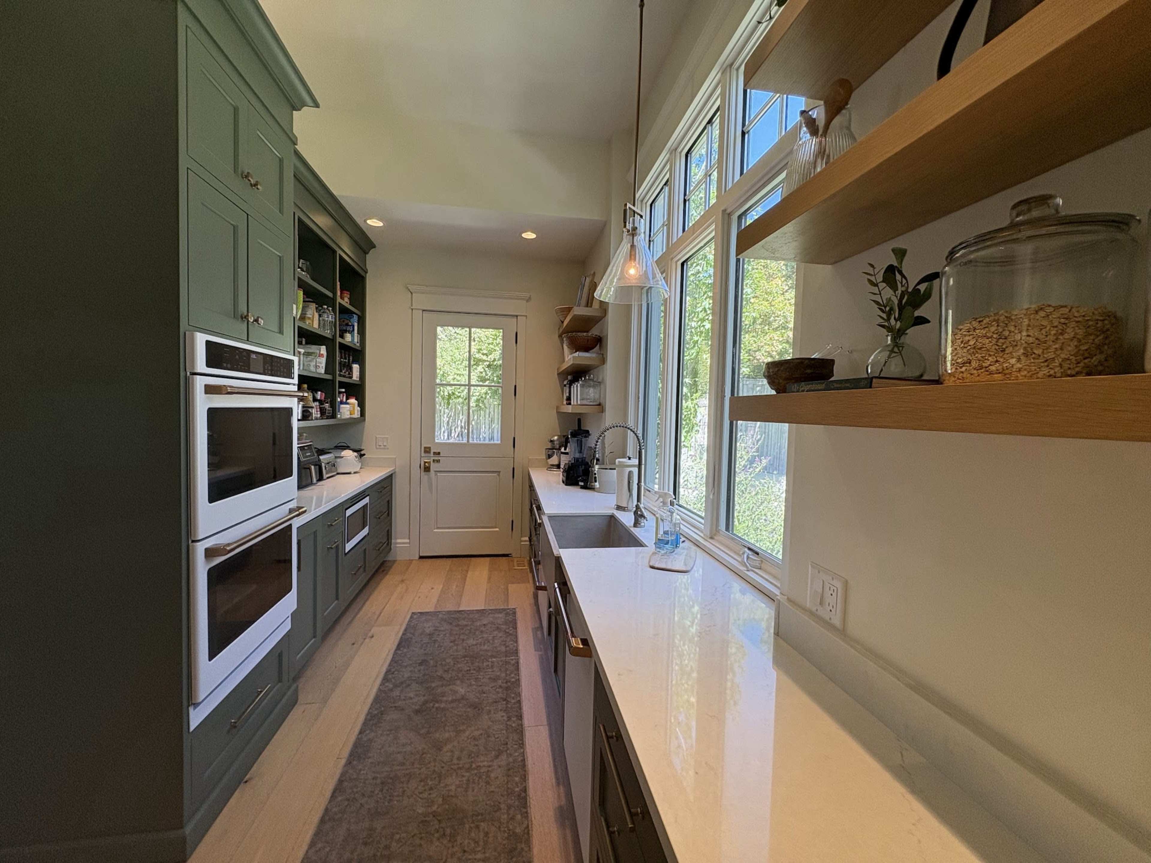 The image shows a modern kitchen with dark green cabinets, white countertops, and large windows allowing natural light to enter, along with shelves displaying various items.